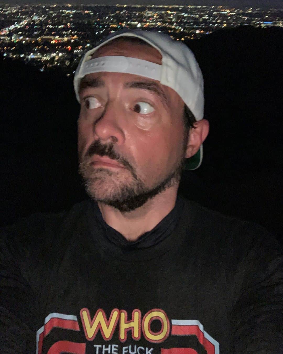 ケヴィン・スミスのインスタグラム：「I was hiking @runyoncanyon last night, and on the benches at the base, I saw a teenager wearing a #Clerks hat, watching a video on his phone. Now I’m not sure if anybody out there knows this, but I *made* Clerks once. So to try to surprise this kid and give him the thrill of his Gen Z life, I stopped and said “I like your hat.” Without ever glancing up, he said “Thanks.” Undaunted, I waited as the kid continued watching his video, lingering for a good minute, hoping he’d eventually make eye contact with his middle-aged stalker and finally recognize Silent Bob. But the kid never looked up. Kids! I’m an old man, so take this advice with a grain of salt. And the advice is this: Look up every once in awhile. The Hat Man might be staring at you. (For those who asked, my t-shirt was made by the genius @stevenrhodesart!) #KevinSmith #runyoncanyon #genz」