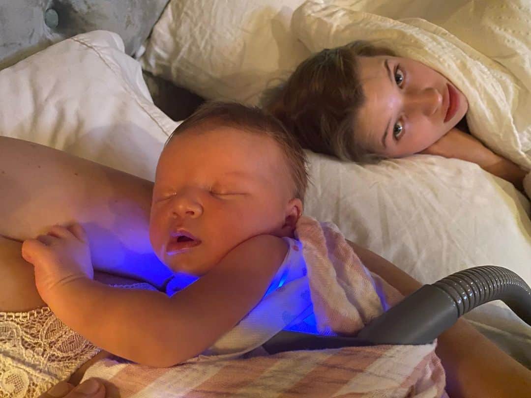 ミラ・ジョヴォヴィッチのインスタグラム：「Poor baby Osian has been having a hard time kicking her jaundice since she was born, but finally we got it under control enough to take her home with a “Bili Blanket” which uses blue light to break down the bilirubin antibodies in her blood. It has a ventilator attached through a tube so it doesn’t overheat which was a bit scary at first, but now I’m used to it i suppose. I wish sunlight was enough, but because of our blood type incompatibility sunlight alone won’t help her😕. Here is a pic that the awesome @chrissbrenner took of what our situation is at the moment. I mean apart from the blue blanket, I could stay like this forever! Laying around in my pj’s all day long nursing a sweet little tiny baby! It’s literally heaven for me. All the time I think that I’m living the best days of my life right now, while these kids are still little! There is nothing better, having them all around us. Spending our days together.❤️❤️❤️」
