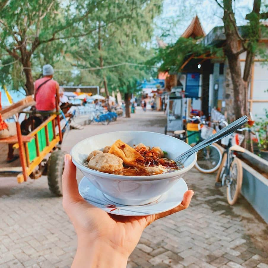 Girleatworldのインスタグラム：「Hi everyone!!! I'm back with a little... 4 year challenge. I managed to find the same Bakso push cart in Gili Trawangan that @singasongjess and I visited 4 years ago! I swear it wasn't planned - I was just biking down the road, spotted a bakso cart and of course I had to stop. It was only after taking the photo and comparing it with the one I took 4 years ago that I realized I was actually standing at the same spot! He was still charging the same amount per bowl. The island has changed quite a bit though. The first image was taken in 2019 and the second image is the one I took in 2015. Swipe right/left and spot the difference.  What is Bakso? Ask any Indonesian about Bakso and i'm sure you would be met with excitement. It's a small bowl of yellow noodles, meatballs and fresh beansprouts in clear beef broth, served with fried shallots, chili and kecap (sweet soy sauce). Bakso is traditionally sold on the street in a small push cart (called "gerobak" in Indonesian) that would go around the neighborhood during supper time and you have to catch the bakso guy at the right time. Because of its small portion, we normally eat it as snacks for supper or midday between meals. Nowadays you can also get them at restaurants for a meal-sized bowl. We got this off a push cart in Gili for less than $2 and proceeded to eat it on the side of the street.  #shotoniphone #iphonexsmax #giliislands #gili #lombok #lombokisland #beach #gilitrawangan #visitlombok #explorelombok #girleatworld #indonesia #bakso」