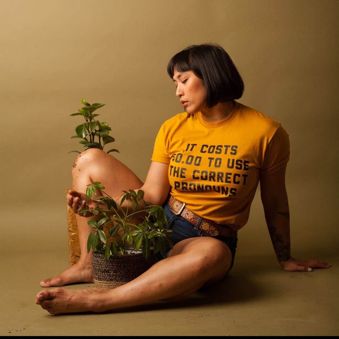 のインスタグラム：「Coming home to myself in the feeling of my fingers in soil. Shot by @taryncarterphoto [photo description: Mia sitting against an olive backdrop. Mia is wearing a gold shirt with the black print saying “It costs $0.00 to use the correct pronouns. There is a plant in a basket sitting between their legs that are curled around it. Mia’s fingers are covered in dirt and she/they are looking at their hand. There is another plant peaking out from behind Mia’s bent knee.] shirt from @greenboxshop」