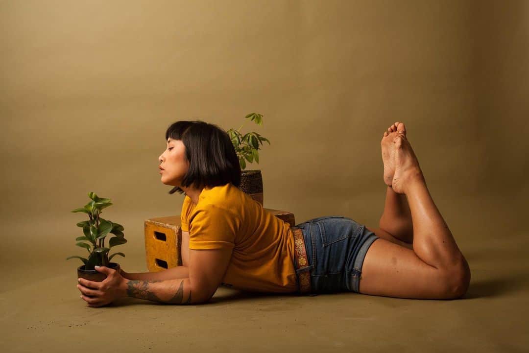 Mia Littleのインスタグラム：「Shot by @taryncarterphoto [photo description: Mia is lying on their stomach propped up on their elbows with their feet kicked up behind them toes touching. Mia is wearing a gold shirt, tan belt, jean shorts with drink on their feet and legs. A plant is peaking behind Mia’s head. They are posed against an olive background.]」