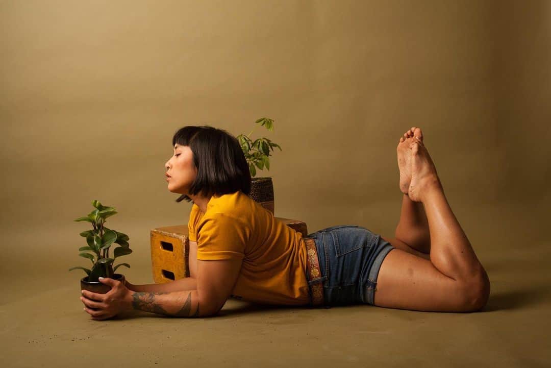 のインスタグラム：「Shot by @taryncarterphoto [photo description: Mia is lying on their stomach propped up on their elbows with their feet kicked up behind them toes touching. Mia is wearing a gold shirt, tan belt, jean shorts with drink on their feet and legs. A plant is peaking behind Mia’s head. They are posed against an olive background.]」