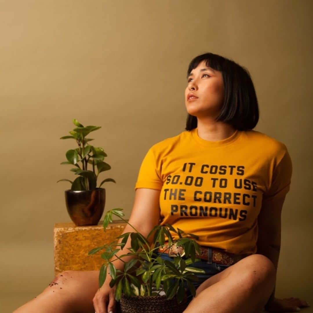のインスタグラム：「Gender is beautiful and can feel like coming home. To share space with those whose embodiment of self reflects back likeness, kinship, and family—it is healing and something I don’t take for granted.  It’s transphobia, racism, and misogyny that feels dangerous, invasive, and violent and is in real ways dangerous, invasive, and violent. Thank you to the community of queer and trans sex worker elders and peers for holding me in spaces, places, and experiences that brought me closer to myself.  Shot by @taryncarter shirt by @greenboxshop [photo description: Mia sitting on the ground with an olive backdrop. Mia is wearing a gold shirt with black text that reads “It costs $0.00 to use correct pronouns” as well as jean shorts. Mia’s legs are smudged with dirt and there is a plant sitting in a basket between her legs and one sitting behind them on an apple box. Mia is looking upward.]」