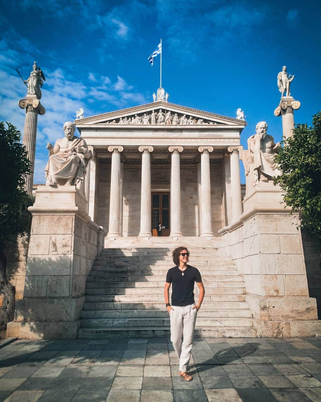 パヴェル・ドローストのインスタグラム：「The Pearl of Athens 📍Academy of Athens 🇬🇷 . . . Одно из моих самых любимых мест в Афинах - Афинская академия наук 🇬🇷 Об этом месте знают не все, так что им можно насладиться в спокойной обстановке.  И пусть само здание было построено не так давно, а именно в конце 19 века, но оно создает прекрасную атмосферу , и словно возвращает нас во времена античности🏛️. Да и к тому же, построена Академия на месте, где в 385г. до н.э Платон также основал свою академию. Именно поэтому, на подступах к Академии нас встречают статуя  Платона и статуя Сократа 💭 По обеим сторонам также высятся статуи богини Афины и Аполлона. #greece #athens #thisisathens #visitgreece #афины #греция #travel #history #vacation」