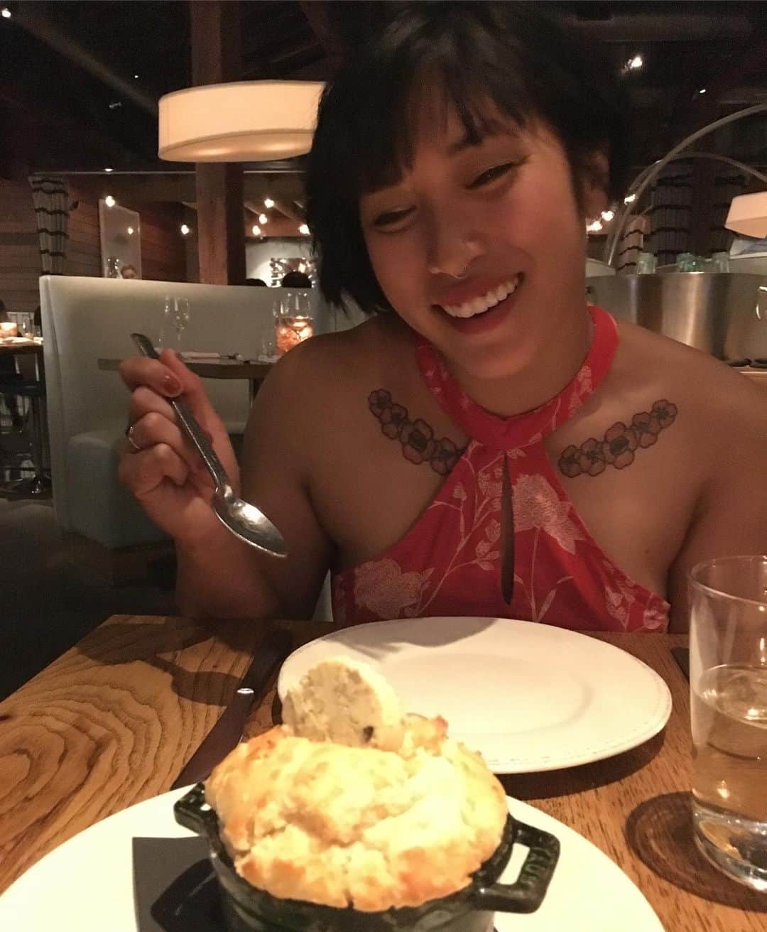 のインスタグラム：「Captured by my best friend. [photo description: Mia looking longingly with a hand holding a raised spoon at a biscuit in a mini cast iron pot topped with a disk of butter. Mia is wearing a red halter type dress that is red with a white floral pattern. The picture is dimly lit and there are restaurant lights and seating in the background.]」