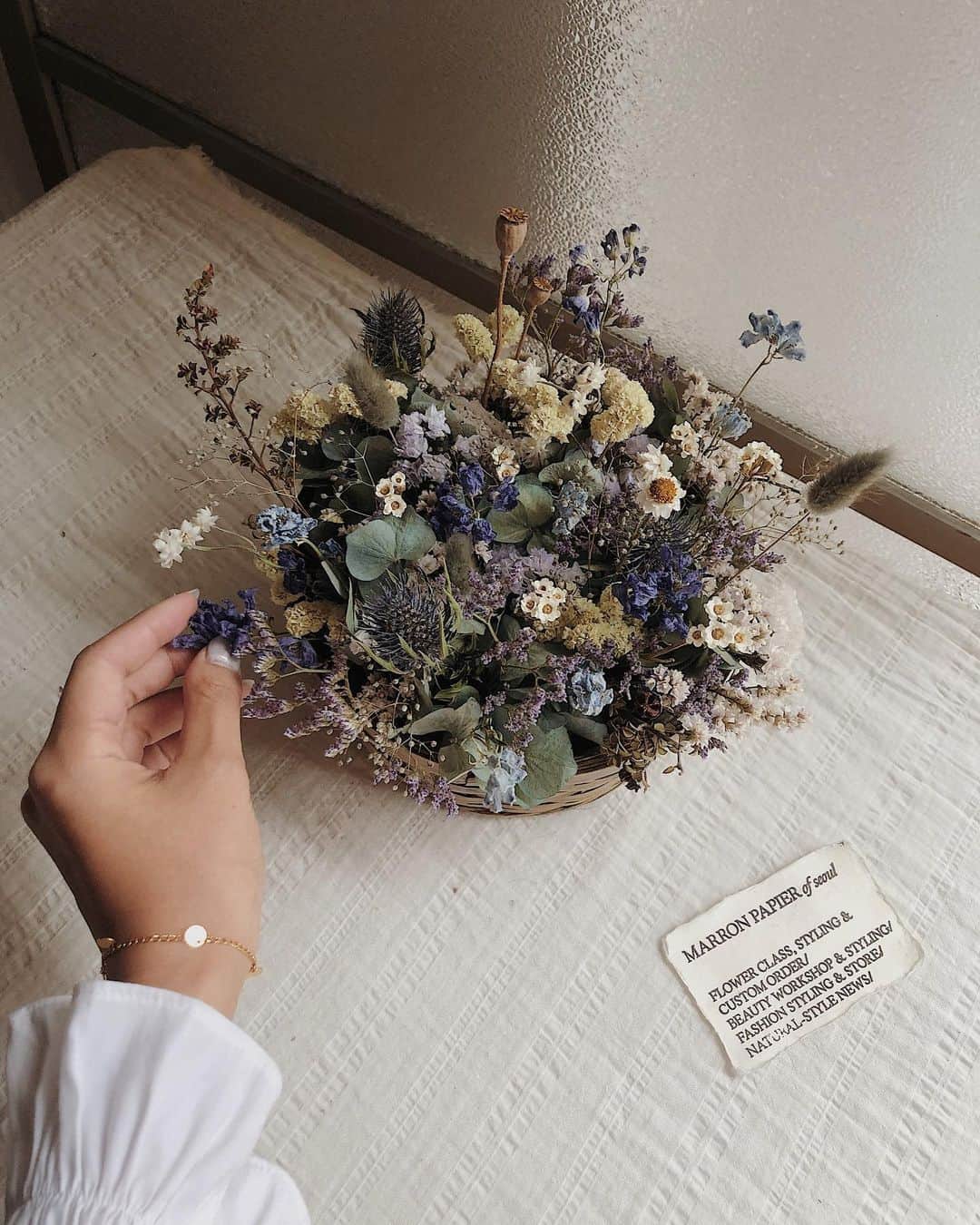 目見田早弥子のインスタグラム：「| Flower Lesson𓆸  前回のマロンのレッスンは ドライフラワーのバケットアレンジメント🧺 日本やヨーロッパのアレンジメントは 真ん中を高く、周りを低くして ドームのように形を作るのが主流だけど マロンのアレンジメントはその逆で 真ん中を低く、周りを高くして 遠くから見た時により奥行を感じるものが ベーシックだそう。 うまくバランスを取りながら周りに高さが出るようにするのがなかなか難しいけど楽しかった！ 私の場合、一周ぐるっと周りを高くしようとすると うるさくなると思ったので緩急をつけるために 一部分だけスターチスやシネンシスをたくさん使い 他では高さを出したデルフィニウムなども低めに、 ザワザワ息詰まらないよう爽やかにまとめました。 ドライよりも生花が好きだけれど このバケットアレンジメントはすごくお気に入り！ 娘も気に入ったようでリビングに飾ったところ、 お花を眺めたりちょんちょん触ったりしています🌼 ラグラスを猫だと思っているようで 指をさして「にゃんにゃん！」と何度も😂🐈 次のレッスンはかなり先なので寂しいなぁ☁️ 先生、いつも楽しい時間をありがとうございます💞」