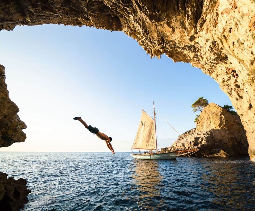 National Geographic Travelさんのインスタグラム写真 - (National Geographic TravelInstagram)「Photo by @Chrisburkard | We came to Mallorca for the seaside climbs and could have stayed for years exploring new ones each day. The island’s perimeter is almost entirely made up of limestone cliffs like these and is always paired with this bright blue water. Our primary mode of transportation was sailboat, as pictured. The climbs were often done without rope, a style of climbing called "deep water soloing." In this style, the water below acts as your safety device in the case of a fall. But, even if you complete your climb successfully, you still have to make it back to the boat. If you look closely, you can see this diver is still wearing his climbing shoes as he plunges into the ocean to swim back to the home base. Not a bad commute.」7月14日 4時01分 - natgeotravel