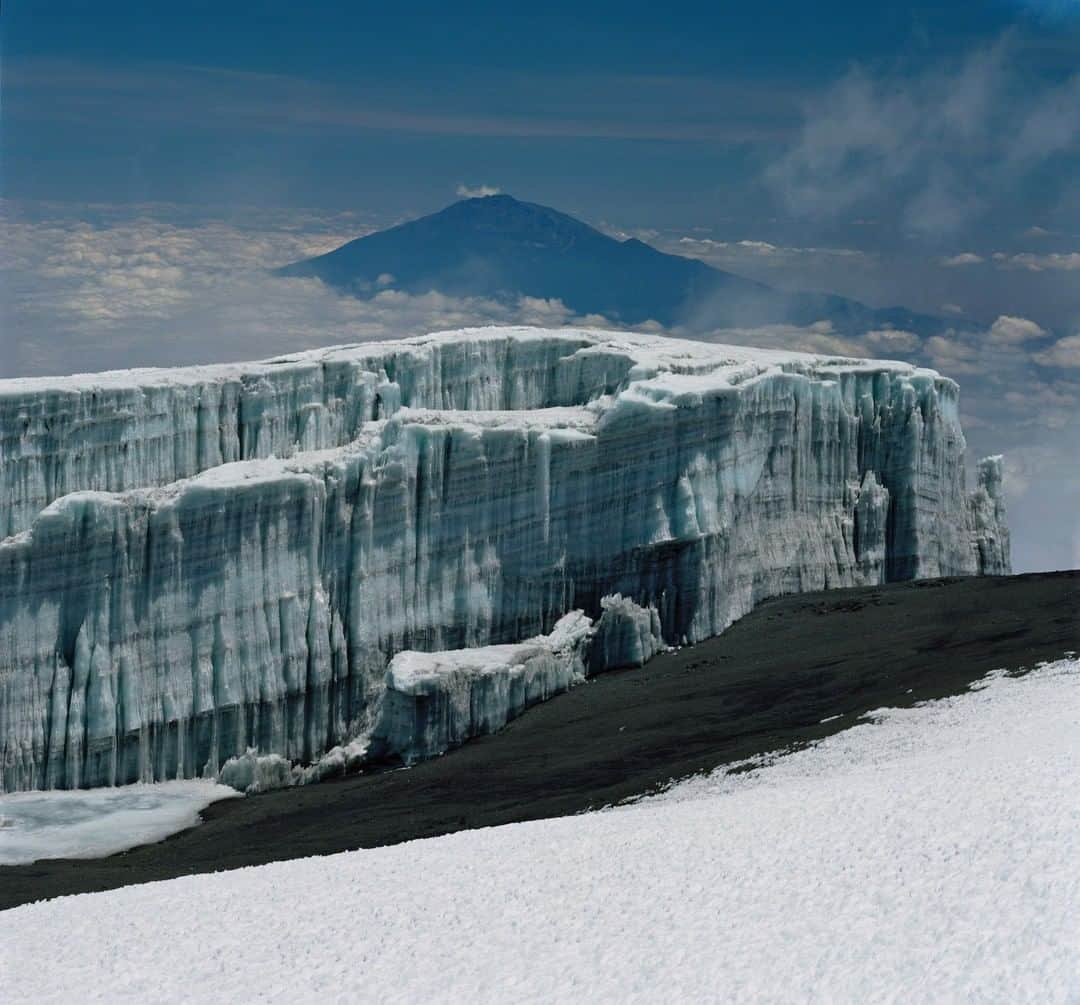 National Geographic Travelさんのインスタグラム写真 - (National Geographic TravelInstagram)「Photo by Simon Norfolk @simonnorfolkstudio I The Kersten Glacier margin, just below Uhuru Peak, with Mount Meru in the distance, photographed whilst climbing Mount Kilimanjaro, Tanzania. Despite continuing shrinkage of Kilimanjaro's glaciers, the steep margins of the Kersten Glacier remain impressive—the cliff is approximately 18 m. (60 ft.) high. The dust bands in the ice absorb more solar radiation than the surrounding, relatively-clean ice, and absorbed energy leads to melting and sublimation. Such stratigraphy is testimony to the complex ice history on Kilimanjaro. Follow me, @simonnorfolkstudiofor, for updates, outtakes, and unpublished and archive material on this and future projects. #photojournalism #Kilimanjaro #climatechange #glaciers」7月3日 7時01分 - natgeotravel