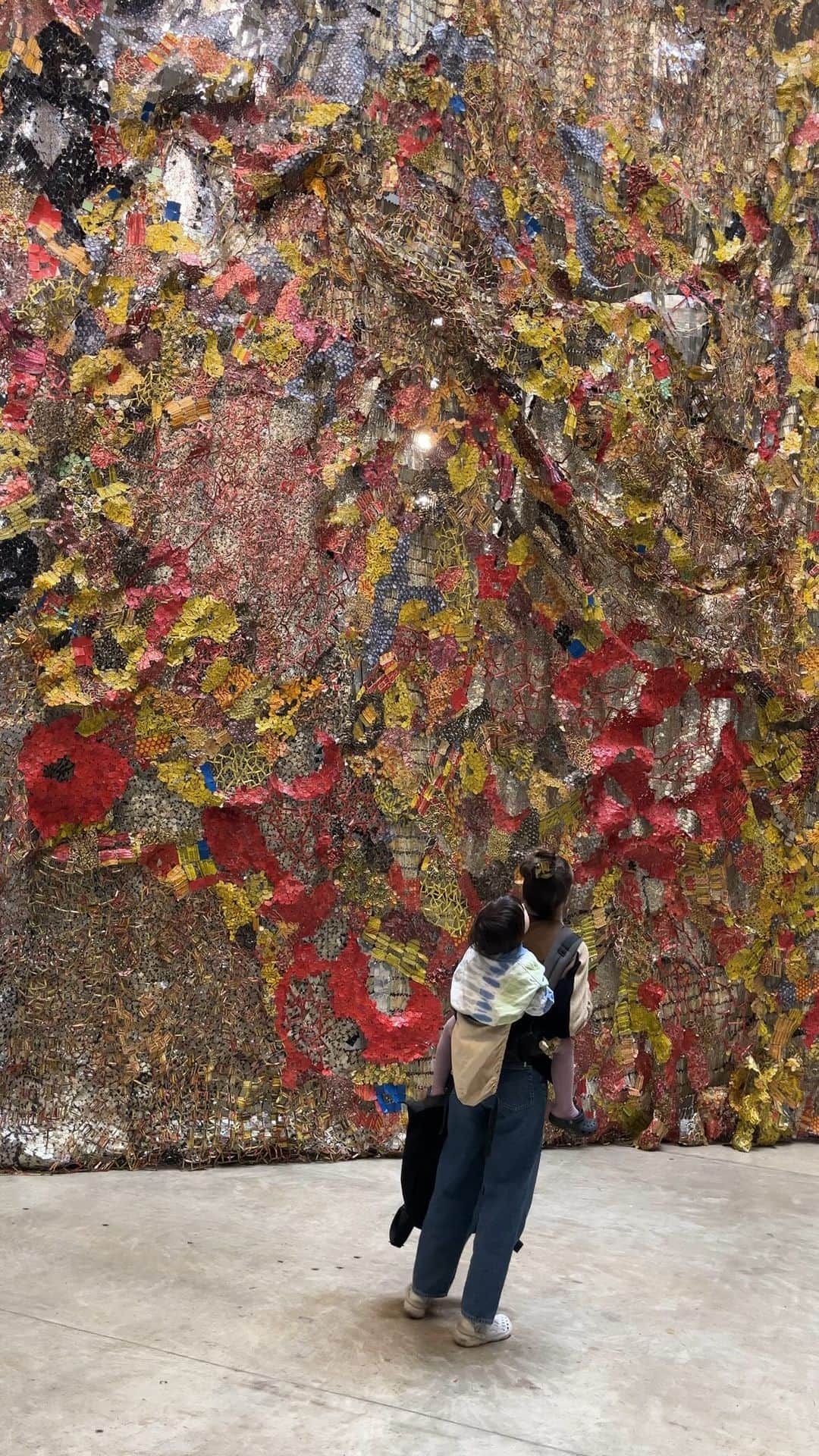 のインスタグラム：「Step into the #TurbineHall and let your gaze be lifted upwards - El Anatsui’s monumental #BehindTheRedMoon sculpture invites you to embark on a three-part journey of light, colour and movement. ✨ 🌙  Up close, this large scale installation made of bottle tops tell stories of global connections, the alcohol trade, and African-European histories. From a distance, the clinking metals form landscapes - waves, moon, earth and sky. 🎨 On display until April 2024. @elanatsui_art」