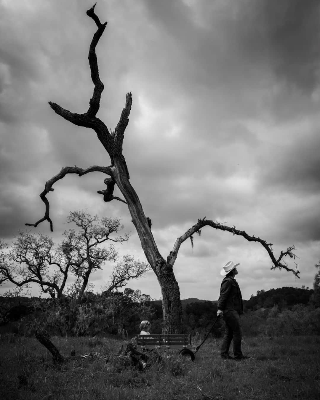 ジョシュ・ブローリンさんのインスタグラム写真 - (ジョシュ・ブローリンInstagram)「When I look at the fence up by where the chapel will be I see that the oak tree that stands in for a corner post has grown around the barbed wire like a freeze frame of a pockmarked landslide. You want to put your little hand on it, attracted, I know, to its unique topography, its gnome like suggestions. You take your tiny steps through waist high oat straw, squinting as you pass the little feathered arrows topping them so to protect your eyes. You look back at me to make sure I’m still there before you place your hand flat against the tree and where it’s built up, where the wire lives underneath. You leave your hand for a moment while I look at the back of your head and notice how silken your hair is, how long it’s gotten. I’m also trying to imagine your thoughts so you don’t do anything rash, like wrap your hand around the rusted barbed wire to the left and right; I’m here to protect you, a job that’s born into me as purely as it is to breathe. But neither of us move. All that sounds are woodpeckers knocking their way into other trees, and a distant cow calling for her calf. When I was your age there were thick mugs being thrown through windows, and drunken nights when my mother would pull men across tables and kiss them while their legs dangled. There were 100 mile per hour drives home, and there were cowboys who hoisted themselves onto 18 wheelers to shut out the screeching nag of my mother’s voice. The way you stand so still with your hand on the tree and the sharp wire underneath as we listen to the return call of the lost calf somewhere on the next ranch over reminds me of when she was still here. You, like white against black or black against white, remind me of her. —  Photo by @kathrynbrolin」6月25日 2時33分 - joshbrolin