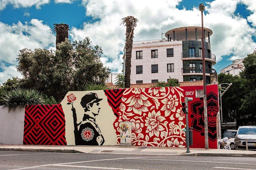 Shepard Faireyのインスタグラム：「The crew and I went to Lisbon, Portugal in 2017, where I opened a show at @Underdogs_Gallery and painted three murals, including a collaboration with @Vhils - one of my favorite artists and the owner of the Underdogs Gallery. For anyone who's never been there, Lisbon is an extraordinarily beautiful city. The architecture, the cobblestone streets, the ceramic tiles, and the wrought-iron balconies have more than enough charm by themselves to be a great reason to visit Lisbon. When you add the contemporary murals, many of which have been facilitated by the Underdogs crew, Lisbon is a must-see for any connoisseur of aesthetics throughout the ages. We were lucky to be met with great weather, and the murals came together pretty quickly thanks to the help from my hardworking crew.⁠⠀ ⁠⠀ My collaborative mural with Vhils was a very fun, organic process, where I painted my half of the mural and some background elements for his half, and he then chiseled his portrait including a small amount of overlap with my image. I love Vhils’s work because of the texture and depth but also because I like the idea of creation and destruction occurring simultaneously. Our mural features two women merging who are from different cultural backgrounds but have a common humanity. I think the aesthetics and the concept of the mural came together powerfully and I’m proud and honored to collaborate with Vhils.⁠⠀ ⁠⠀ I also painted two other murals where I used the motif of the flower in the gun, a peace symbol inspired by war protestors putting flowers in the barrels of the National Guards guns during the Vietnam War era protests. I was happy that a symbolic motif that I’ve used in my work had a historical connection to the 1974 bloodless Carnation Revolution in Portugal, which ended a dictatorship and established democracy.⁠⠀ ⁠⠀ The show at Underdogs was nice and crowded, and I really enjoyed meeting so many friendly and enthusiastic people during the opening. The Underdogs crew did an amazing job producing the art show and facilitating the mural walls. Thanks to Vhils & the whole Underdogs family. I look forward to visiting Portugal again someday!⁠⠀ – S⁠⠀ ⁠⠀ Full recap through the link in bio!」