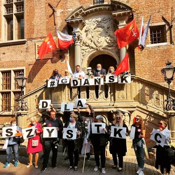 ポーランド政府観光局 ZOPOT w Tokioのインスタグラム：「さて、こちらは今日グダンスクの旧市庁舎前での一枚。皆さんが持っているのは「みんなのためのグダンスク」というスローガンです。この旧市庁舎は旧市街の中心、半キロ近く続く通りul. Długa ( ドゥーガ通り)にあります。じつは通りが町の中心の広場になっているのはなかなかレアなのです。中世からあらゆる意味でグダンスクの人々の生活の中心になって来たフォトジェニックな通り。ここに来たら階段の上にのぼって記念撮影をお忘れなく。階段の上からも素敵な写真が撮れますヨ。😉」