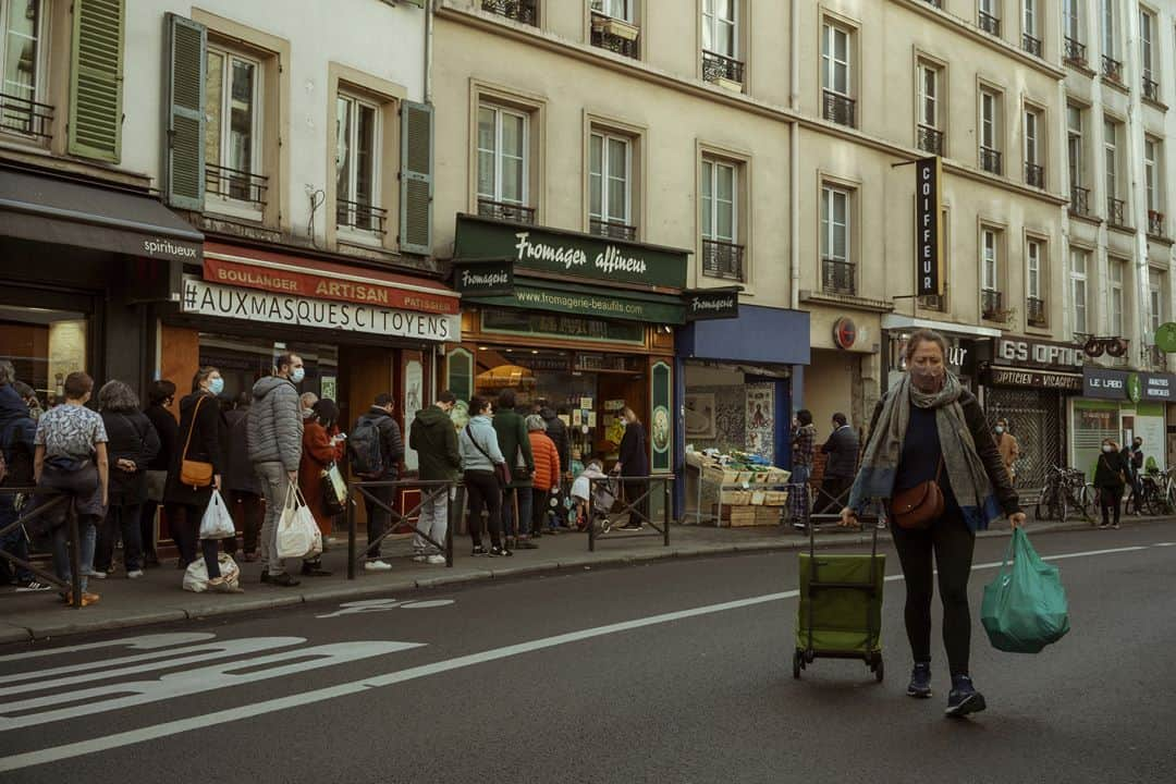 ルモンドさんのインスタグラム写真 - (ルモンドInstagram)「La rue de Belleville, à Paris, accueille paresseusement ses habitués du samedi matin. Au bar des Folies, les réguliers sont à peine perturbés par la transformation de l’institution du quartier en épicerie-pizzeria. « On est des caméléons. On regarde tous les jours ce qu’on a le droit de faire, et on s’adapte », s’amuse presque le serveur. Ce n’est pas pour déplaire à Khalil Ghabara, qui continue à venir tous les matins, « comme avant ». Il sort plusieurs fois par jour, usant d’une attestation puis d’une autre. « Mais toujours en règle ! »⁣ Les stores baissés se font de plus en plus rares en remontant la rue, et les trottoirs se gorgent de badauds. Un couple s’arrête devant une papeterie, ouverte elle aussi. « Dis, chéri, on ne rentrerait pas là ? On n’y est jamais allés ! » Boulanger, boucher, poissonnier, fromager… Les files s’étirent, moins longues que lors du premier confinement mais pleines toute la journée. « Est-ce que c’est très raisonnable ? Je ne sais pas », convient Sabine Civilise en attendant son tour – « J’avais envie de lapin ! » – à la boucherie.⁣ La boutique vide et l’air contrarié, Mahnaz la chocolatière observe la rue débordant de vie en soupirant. « Les gens sont des enfants, les policiers sont occupés ailleurs, alors, ils en profitent pour passer entre les gouttes… » En Iran, Mahnaz a connu la guerre, puis l’embargo. « Du coup quand les gens disent : “J’en ai marre d’être confiné”, alors que les rayons des magasins sont pleins, je préfère ne rien dire… » Sans en penser moins.⁣ -⁣ 1 : Le café des Folies s’est reconverti momentanément en pizzeria-traiteur pour contourner les mesures de fermeture⁣ 2 : Foued, peintre, boit un café rue de Belleville, le 7 novembre⁣ 3 : Les files d’attentes devant les commerces de bouche de la rue de Belleville⁣ 4 : Davy et Stéphane, après avoir fait la fête toute la nuit chez des amis, sont à la recherche d’huîtres et de vin blanc sur le marché⁣ 5 : Mahnaz, dans sa boutique de chocolats⁣ 6 : Sophie et Brigitte partagent leur café du matin devant l’église de Belleville⁣ 7 : La rue de Belleville à Paris le 7 novembre⁣ -⁣ Photos : Agnès Dherbeys (@agnes_dherbeys) #PourLeMonde⁣ -⁣ #covid #coronavirus #commerce」11月11日 22時42分 - lemondefr