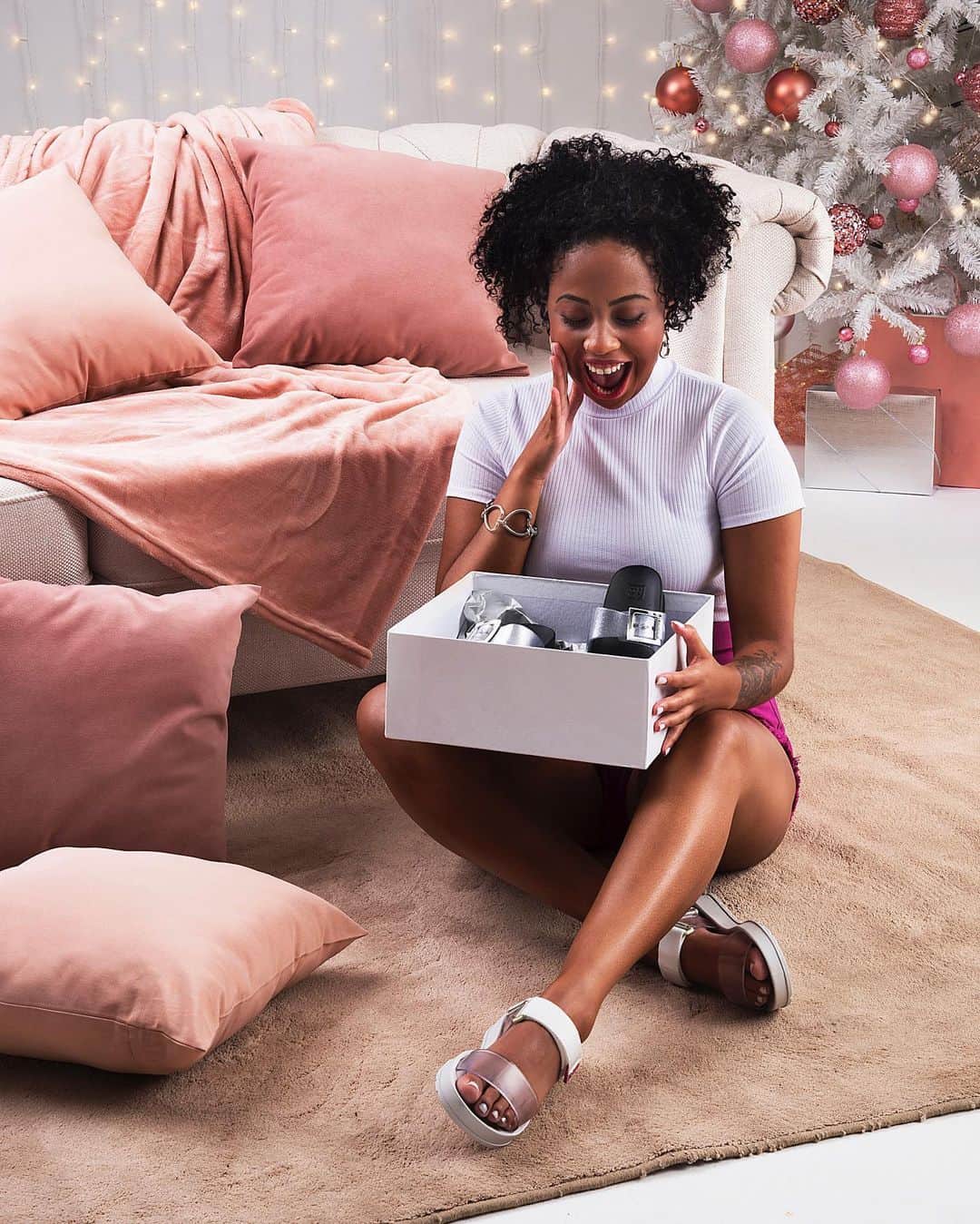 のインスタグラム：「Desejo de Natal: abrir a caixa de presente e encontrar aquela Zaxy que você queria muito 😜 Marca aquela pessoa que vai te dar um presente! #FimDeAnoZaxy  #ZaxyPartner - R$54,99 #ZaxyWayPapete - R$69,99  Compre agora mesmo pelas tags no post, pelo link na bio ou na nossa loja online www.zaxy.com.br  #pracegover #pratodosverem: Na foto, temos uma parede branca ao fundo com luzinhas penduradas. À direita, uma árvore de Natal branca com vários enfeites em tons rosa e rosé. À frente, do lado esquerdo, temos um sofá branco com duas almofadas. No chão, temos um tapete marrom claro. Nele, temos uma mulher negra sentada com as pernas cruzadas. Ela segura uma caixa branca com a Zaxy Partner prata dentro. Nos pés, ela usa a Zaxy Way Papete. A Zaxy Partner possui 2 tiras frontais com fivelas na lateral. A Zaxy Way Papete é uma sandália com duas tiras frontais e o fechamento traseiro.」