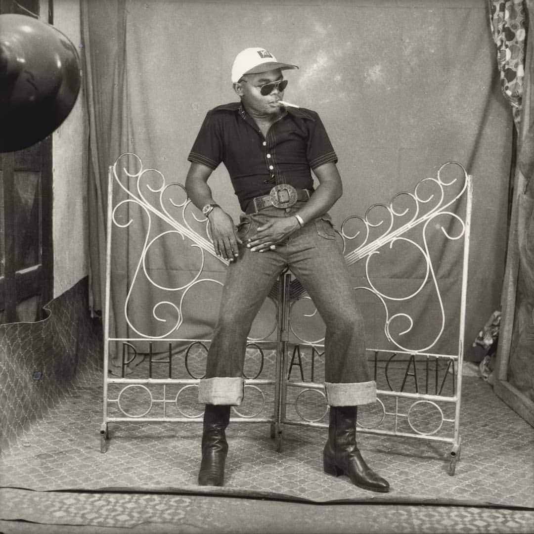 ニューヨーク近代美術館のインスタグラム：「Cool factor 💯 ​ ​Studio portrait photography flourished across Africa in the 1960s and '70s, becoming a popular medium for creative and self expression. ​ This photograph by Sanlé Sory reflects the popularity of American cinema in newly independent Burkina Faso. The James Dean-esque young man leans against an iron gate, perhaps a cheeky interpretation of the white picket fence. He sports the accoutrements of 1950s Hollywood: a baseball cap, aviator sunglasses, blue jeans with an oversized belt buckle, and a cigarette hangs rebelliously from his mouth.  At the link in our bio, explore how photo studios provided collaborative spaces for play and performance through the works of #SanleSory and #AmbroiseNgaimoko, new to the #MoMACollection, and see more of their portraits in our Stories.  Photo: Sanlé Sory, "L’Americain (American)," 1970-85. © 2020 Sanlé Sory, courtesy @yossimilo」