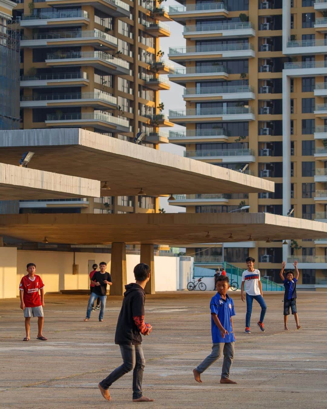 Shunsuke Miyatakeのインスタグラム：「Barefooted children and young men in sandals and shoes enjoy playing football with a Sepak Takraw ball against the backdrop of a modern building under construction in the corner of the Olympic Stadium built by the famous Cambodian architect Vann Molyvann.」