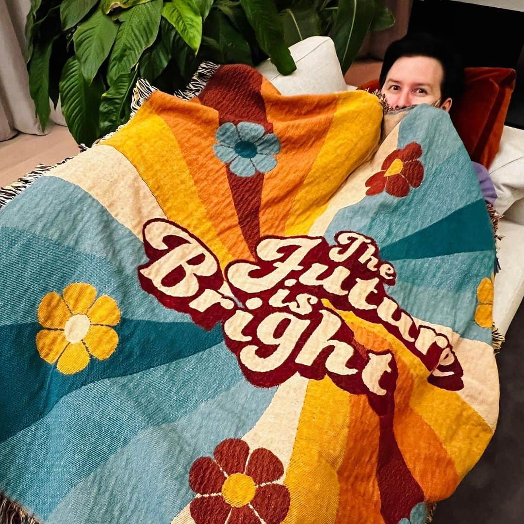 フィル・レスターのインスタグラム：「If you need me I will be snug as a very long bug under this rug until further notice   NEW MERCH!! Prepare to be almost too cozy under my new limited edition ‘future is bright’ handmade woven blanket! AND the most delicious smelling candle on earth - chocolate and toffee ‘treat yourself’ made in collab with @ohroslondon   Available now with loads of other stuff on sale at amazingphilshop.com and us.amazingphilshop.com (combined with Dan shop and danandphilshop for the festive season!)🕯✨」