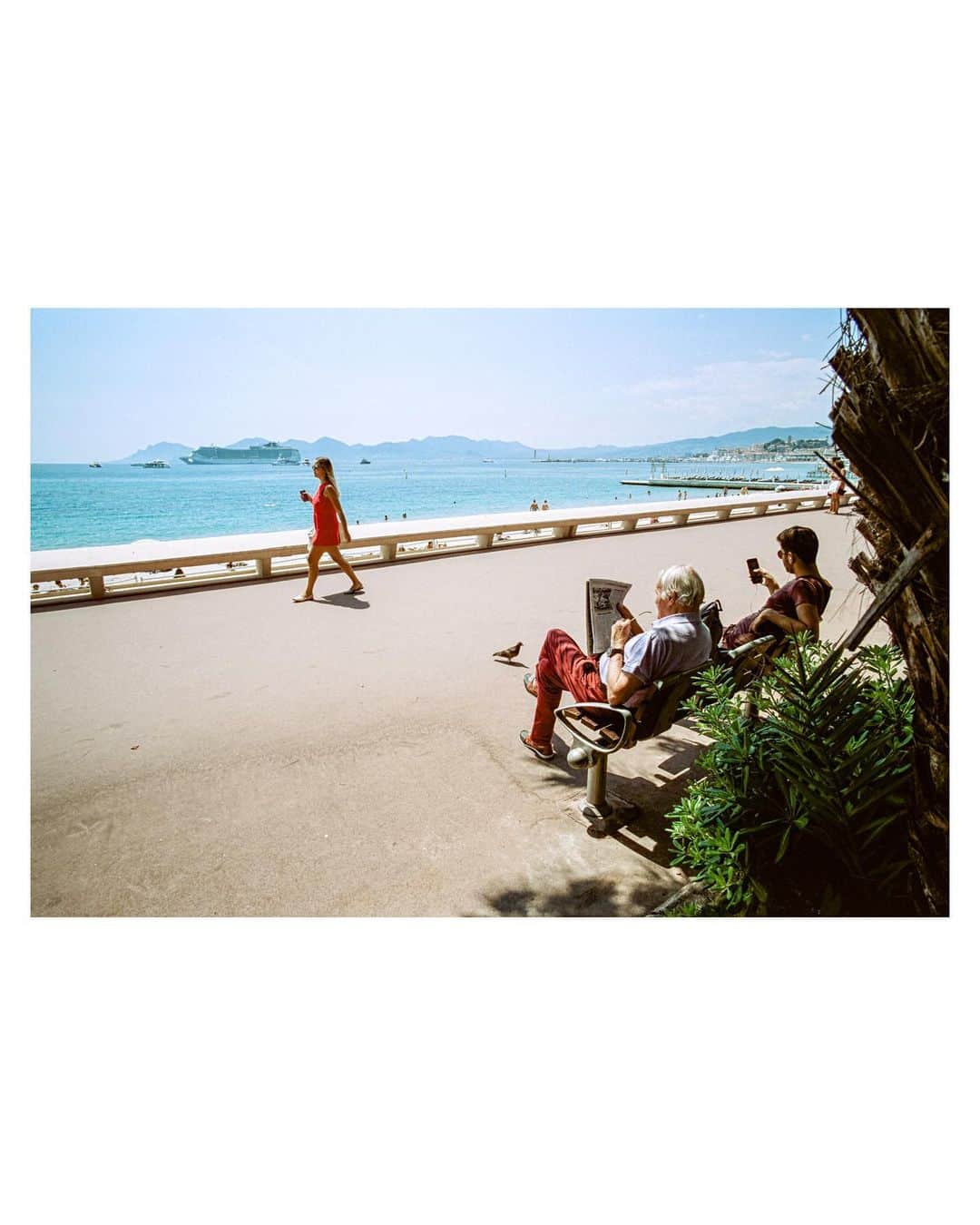 Pete Halvorsenのインスタグラム：「The 3rd image from my Cannes series is now minted and live on @superrare.co   With this image we are back on the world famous Promenade de la Croisette. A short story on this image, and what I look for when shooting on the street:  The scene - I noticed this elderly gentleman sitting reading his paper facing the sea...a young man next to him earphones on lost within his phone. Struck by the juxtaposition of the two generations consuming their information differently on the same bench, I composed my frame.  A Lady In Red approached, both generations of "watchers" decided to look up simultaneously and watch her stroll by. This is where the compose and wait technique pays off...  The pieces of the puzzle are all laid out, it's my job as the storyteller to put them together for the viewer, so without a caption - can the story can still be told?  Can the frame speak for itself?   Colour for me is a big part of that story.  Shooting on film, while more difficult to "quickly review" images creates creative guardrails that allow for the photographer to work within. @cinestillfilm 50D is one of those that compliment my love of bright blues/deep reds...it pairs nicely with a day walking around Cannes.  This is where compose and wait comes in, I had his red pants on one side, just needed something to connect it to. The woman in the dress approached...gave me colour connection for the viewers eye to go back and forth between the two sides of the frame.  Colour...composition...patience, and a bit of luck. Proud to add the 3rd image from a summers day in Cannes...through the eye of my @leica_camera M7 rangefinder, the light frozen in time on CineStill Film and now minted on the blockchain and available as a 1/1 on @superrare.co」