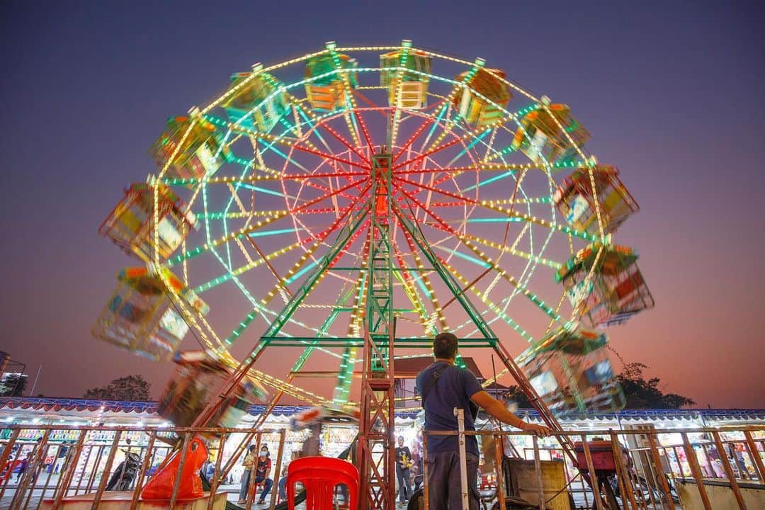 Shunsuke Miyatakeのインスタグラム：「Ferris Wheel in Cambodia /  The man in charge of the Ferris wheel presses a button, the engine starts, and the Ferris wheel slowly begins to spin. The Ferris wheel goes around with a variety of visitors, from small children to adults.  Some people cheer, some scream, and many other voices can be heard.  I wonder how many people have ridden this Ferris wheel. The neon lights shining at dusk were very beautiful. The man in charge pushed a button and the Ferris wheel quietly stopped. The man's back looked a little sad.」