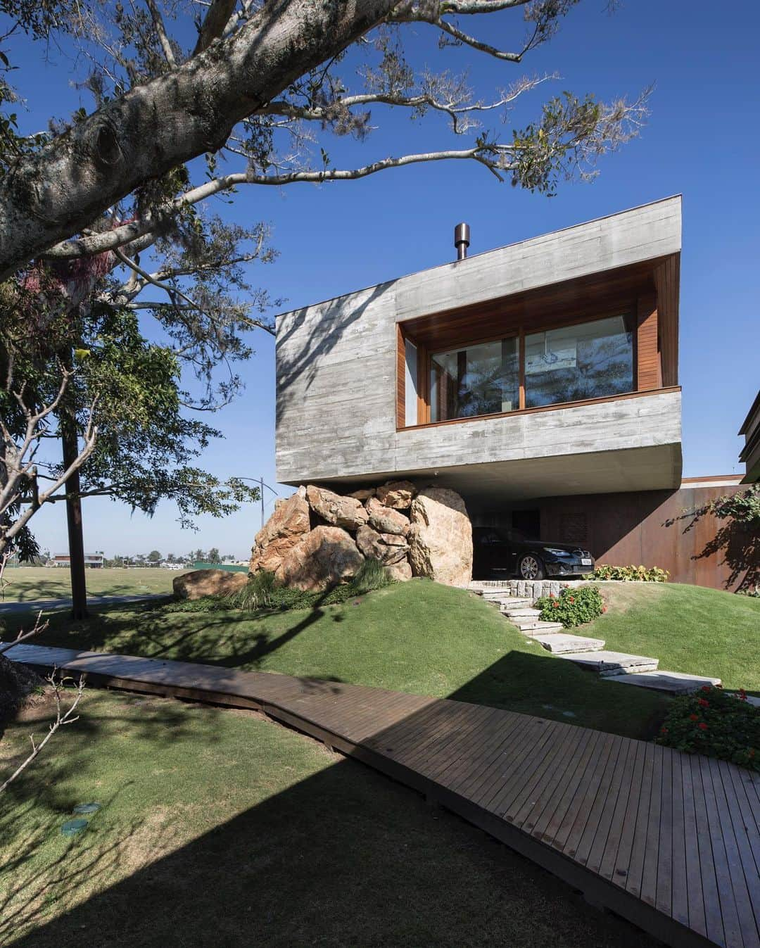 Inspirationのインスタグラム：「A Casa da Figueira é uma residência projetada pelo escritório (@stemmerrodrigues), localizada em Porto Alegre, RS, com uma área construída de 726 m². Situada em um terreno plano, a casa é nomeada em homenagem a uma escultural figueira que é refletida nas águas do canal navegável próximo.  A fachada da casa é composta por uma ampla estrutura de concreto aparente e madeira, que serve como proteção contra a corrente de ar minuano comum na região Sul. O espaço aberto localizado na parte de trás da residência se integra harmoniosamente à natureza, proporcionando uma vista encantadora para o canal.  Uma edícula localizada meio lance abaixo do terreno foi projetada para ser uma área de lazer e para admirar a casa principal. Já no pavimento inferior, localiza-se a área social, com um extenso volume de vidros que permite o contato visual com a ampla vegetação e o canal navegável, elementos marcantes do projeto.  No pavimento superior encontram-se os dormitórios, compostos por caixilhos de vidro que os ligam externamente, e o piso de madeira que se estende pelo terraço. Para garantir maior eficiência energética, foram instaladas claraboias móveis motorizadas no térreo e no primeiro andar da casa, que promovem a iluminação natural e a ventilação cruzada, gerando economia de energia elétrica com climatização e iluminação.  A fachada principal da Casa da Figueira é um misto de rigidez da madeira ipê e do concreto aparente, sendo que uma grande rocha parece sustentar a casa, conferindo personalidade e um contraste marcante ao design.  📸 @donadussi」