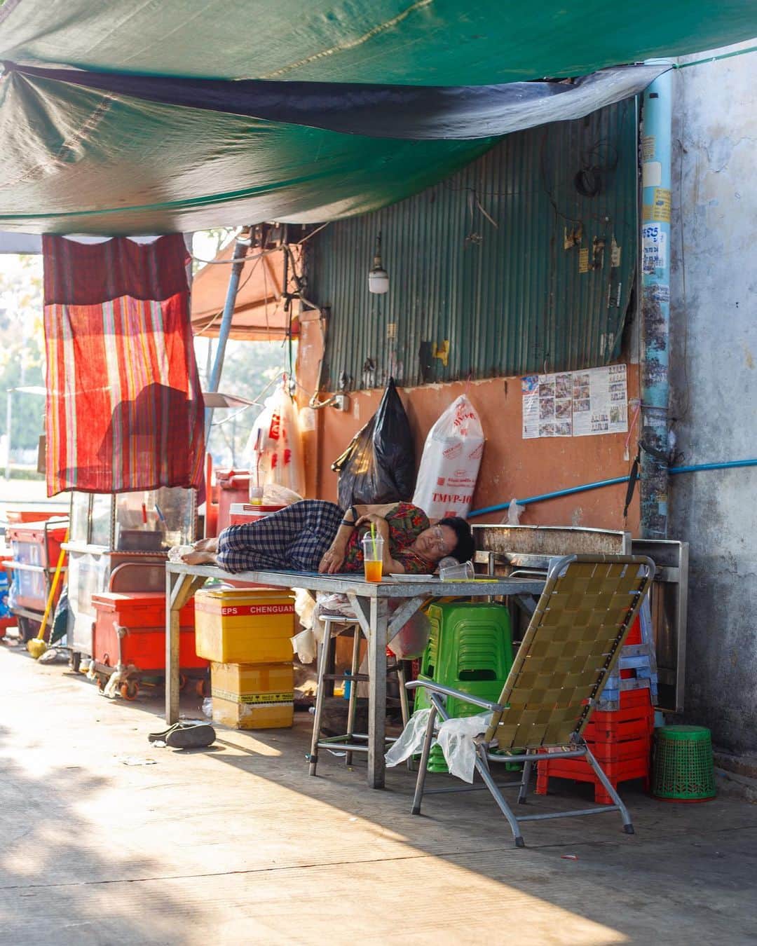 Shunsuke Miyatakeのインスタグラム：「Siesta / 　  Saturday afternoon. It is the time of day when we feel a little sleepy. This woman was lying on a table in a store and sleeping soundly.  During this time of the year, the sun is so strong that we can easily get sweaty when walking outside. Perhaps the shade of the eaves is just right for sleeping.  She sleeps when she wants to, without fear of being seen. Walking around Phnom Penh, I often see people taking a nap.」