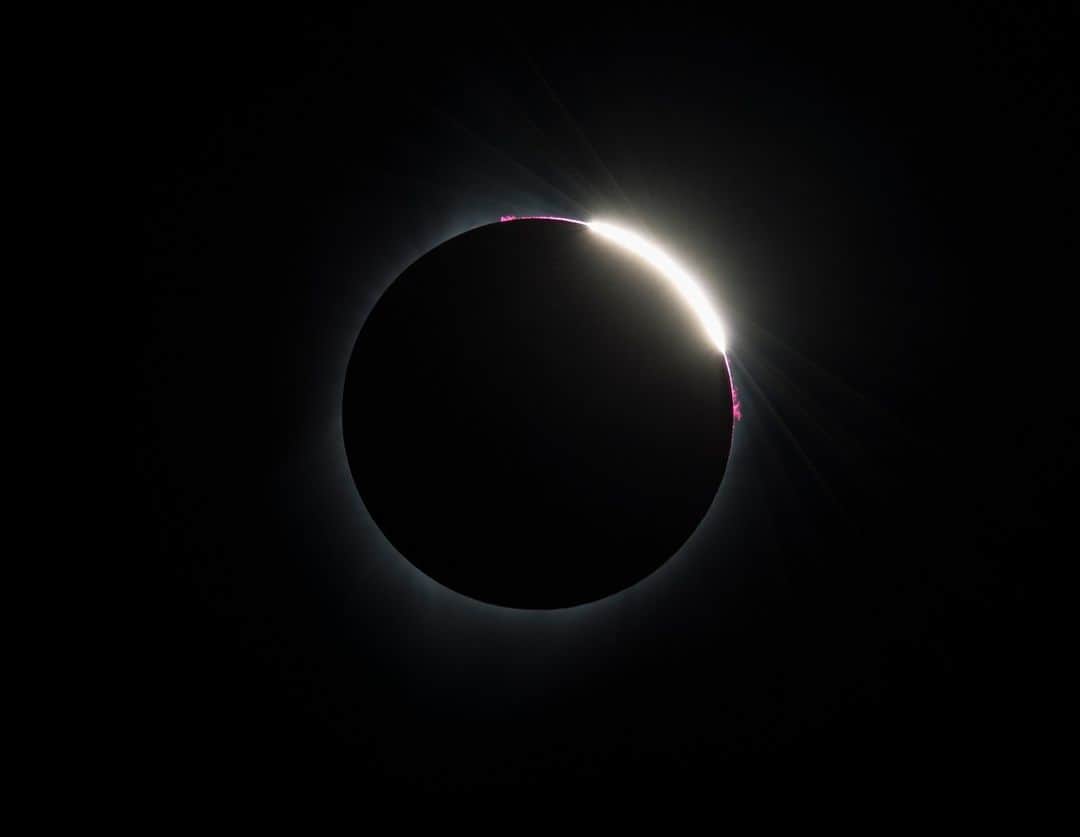 NASAのインスタグラム：「Paint it, black 🖤⁣ ⁣ On April 20, skywatchers in Australia and parts of southeast Asia will be able to view a hybrid solar eclipse as it passes over the Indian and Pacific Oceans—while the rest of the world can follow the links in our bio to check out live streams on NASA's YouTube and Twitch channels! (Links in bio.)⁣ ⁣ Tolar eclipses occur when the Sun, the Moon, and Earth fully or partially align, casting a shadow on our home planet and obscuring the bright face of our closest star. Remember, it is unsafe to look directly at the Sun for most of the eclipse without specialized eye protection.⁣ ⁣ There are several types of eclipses, total, annular, hybrid, and partial. In a total eclipse, the Moon entirely blocks the Sun allowing people in the eclipse's path to see the star's corona – outer atmosphere. Total eclipses are the only type where viewers can momentarily remove their eclipse glasses. In an annular eclipse, the Moon perfectly lines up with the Sun but is farther away from the surface of Earth and does not fully cover the Sun's face. Hybrid eclipses shift from annular to total due to our planet's curve. In partial eclipses, the Moon is not fully lined up with the Sun, only partially covering its bright face.⁣ ⁣ Image description: In the center of the image, a faint white outline of the Sun's corona appears behind the Moon as it is fully obscured during a total eclipse in 2017. On the top corner of the eclipse, a bright yellow-white section sends rays of sunshine in several directions, with a bright pink outline next to it.⁣ ⁣ Credit: NASA/Aubrey Gemignani⁣ ⁣ #NASA #Eclipse #Sun #SolarSystem #SolarEclipse」