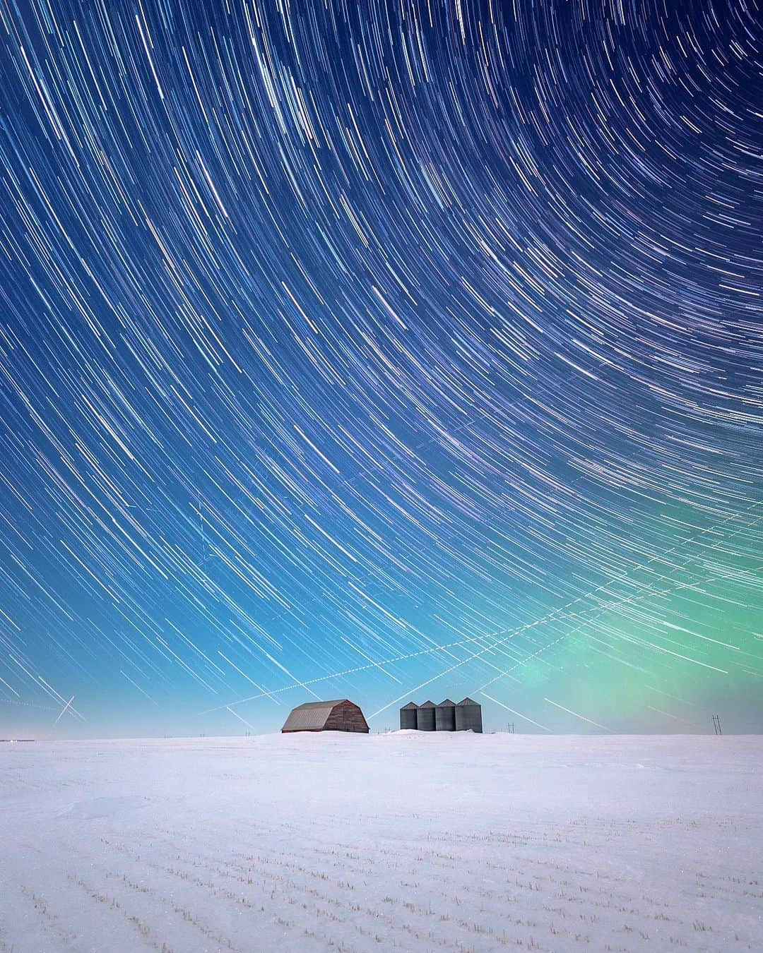 のインスタグラム：「Don’t forget to look up 🌠  📷: @herry.with.an.e  📍: @tourismsask   #ExploreSask  [Image description: A wooden barn and four metal silos in the middle of a snowy field under a night sky. The stars in the sky show light trails, created by a long exposure in the camera.]」