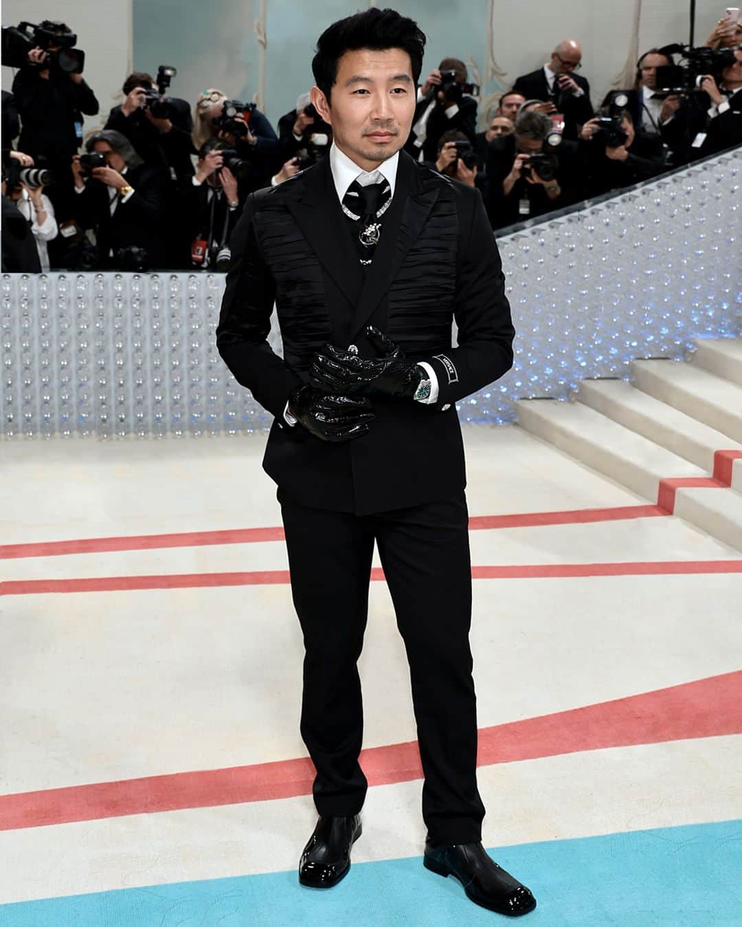 ジャンニヴェルサーチのインスタグラム：「Simu Liu in custom Versace at the MET Gala 2023.  Simu Liu embraces the iconic image of Karl Lagerfeld himself, with a custom Versace suit.  The Grain de Poudre wool single-breasted blazer features a peak lapel, silver Medusa buttons, a Versace tailoring label at the sleeve, and silk satin plissé inserts. It is worn with matching smoking pants with silk side band, and over a shirt with Karl's distinctive high-neck collar, and a custom tiepin. The look is styled with crocodile embossed leather gloves and patent boots.   @simuliu  #AtelierVersace #MetGala #MetGala2023」