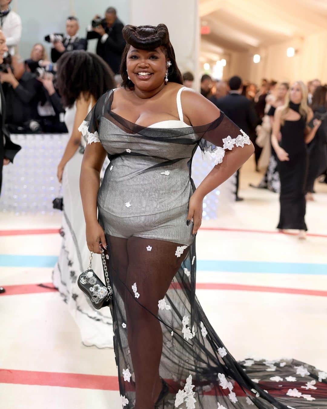 のインスタグラム：「Give it up for Gabby. 🤍 Fashion editor and stylist #GabriellaKarefaJohnson finds her light in a custom @coachtopia gown. Inspired by 1930s-style silhouettes, the outer dress is cut in silk and cotton tulle from remnants of the #CoachFall20 runway collection. The inner portion is comprised of a full corset and knit lace stitch bloomer made from recycled yarn. Hand-appliquéd lace intarsia covers the dress (dyed naturally using dandelion tea 🌼) and the smile is a *Gabriella takes the #MetGala* original. Gabby also carries a Wavy Dinky bag embellished with Met Museum discards—maps, plastic water bottles and napkins.   Photographer: @benjaminrosser Hair: @lacyredway  Nails: @nailglam Makeup: @raisaflowers #CoachNY #Coachtopia」