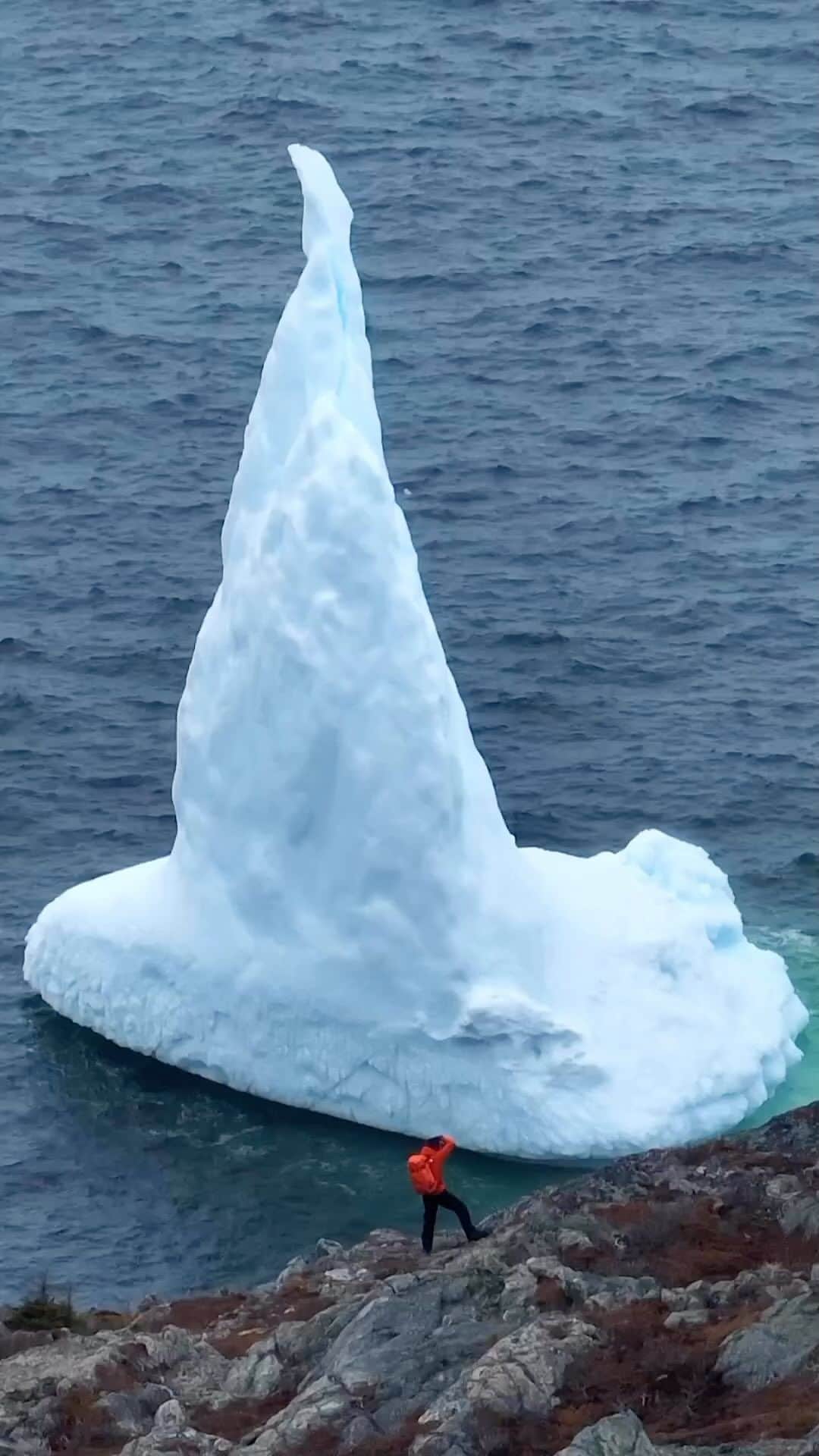 Explore Canadaのインスタグラム：「These guests visit every year!   Did you know that around 90% of the icebergs seen off of the coast of @newfoundlandlabrador come from the glaciers of western Greenland? Their sheer size will amaze you, and that’s not counting the 90% still below the water.   You can watch icebergs from the shore (we recommend binoculars) or join a local boat tour operator for a guided experience.   📷: @tomcochrane 📍: @icebergfinder, @newfoundlandlabrador   #ExploreNL   [Video description: A person wearing an orange coat and backpack stands near the shore taking a picture of a large iceberg floating in the ocean. The bright white iceberg is a tall, triangle shape.]」