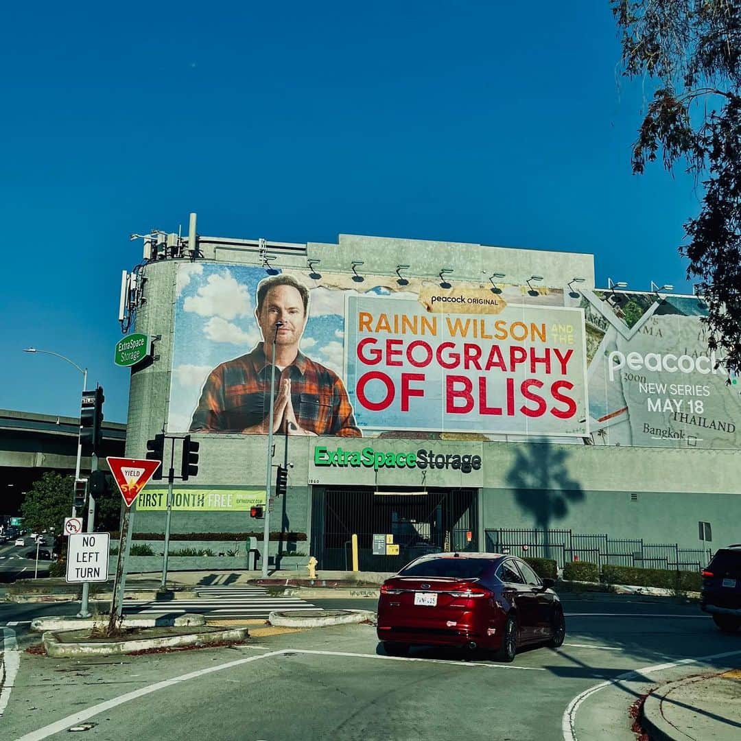 レイン・ウィルソンのインスタグラム：「This sign is right in Hollywood right off of the 101 freeway, about half a mile from my first apartment when we first moved to LA. I paid $650 a month for a one bedroom the size of a shoebox. I had just arrived from New York, where I had only done theater, and I was striking out in the new arenas of television and film in and about the year 2000. That year I did TV minor guest actor roles on Charmed and CSI. I was also teaching acting to foreign students at a hole in the wall school on Hollywood Boulevard that my friend ran. I was in a constant battle to pay my bills, and I was in a constant internal battle… wondering if I would ever really make it in showbiz. Or if I should move back to my hometown of Seattle with @holidayreinhorn and give up on the whole enterprise. Sell salmon or something.  And now? My giant head on the side of a Hollywood self storage facility. Visible to the Rush of cars streaming down the 101 towards downtown. Practically visible from the window of my first apartment in Hollywood all those years ago.  I guess I “made it.” Weird.  So grateful to be a part of this amazing show. A show that actually makes the world a better place. Finding happiness in different cultures and bringing those findings home for you the viewer. And for me the fucked-up host. #GeographyofBliss on  @peacock」