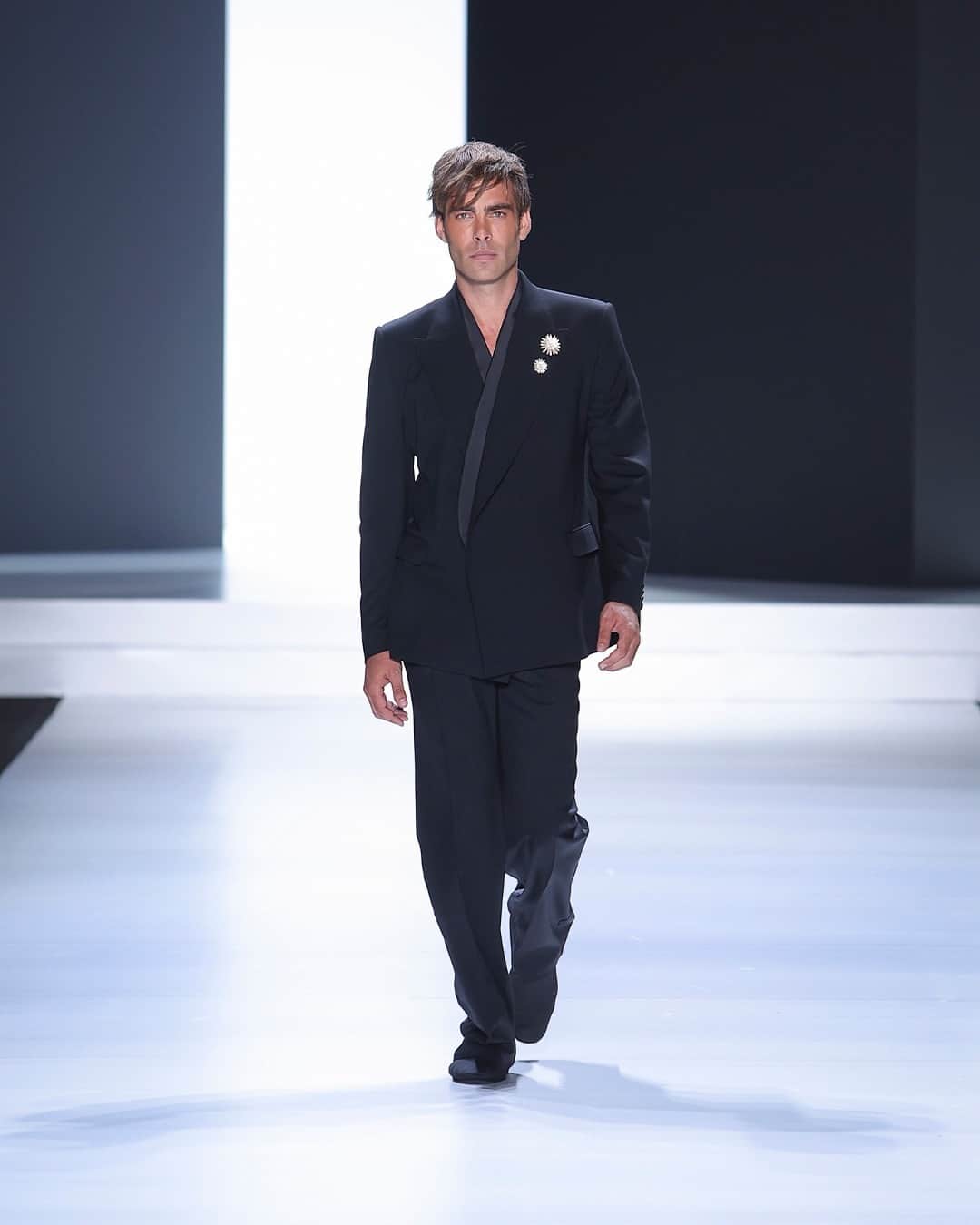 のインスタグラム：「#DGSS24 Men’s fashion show "STILE": enhancing rigor, emphasizing and reaffirming #DolceGabbana original codes.    @jonkortajarena models a Sicilia jacket with a peak lapel and off-centre buttoning. The jacket combines two materials, wool and silk satin, and is adorned with two flower brooch pins. The outfit is completed with plain-front trousers.    @abas.abdirazaq models a complete outfit featuring a striped poplin shirt with long sleeves, paired with cotton drill 40-bottom trousers, a black wool hat and black moccasins.    A tailored coat with a peak lapel made of technical wool, adorned with 3D flower pins. The outfit also includes black Chantilly lace trousers and a top, along with black satin lingerie.」