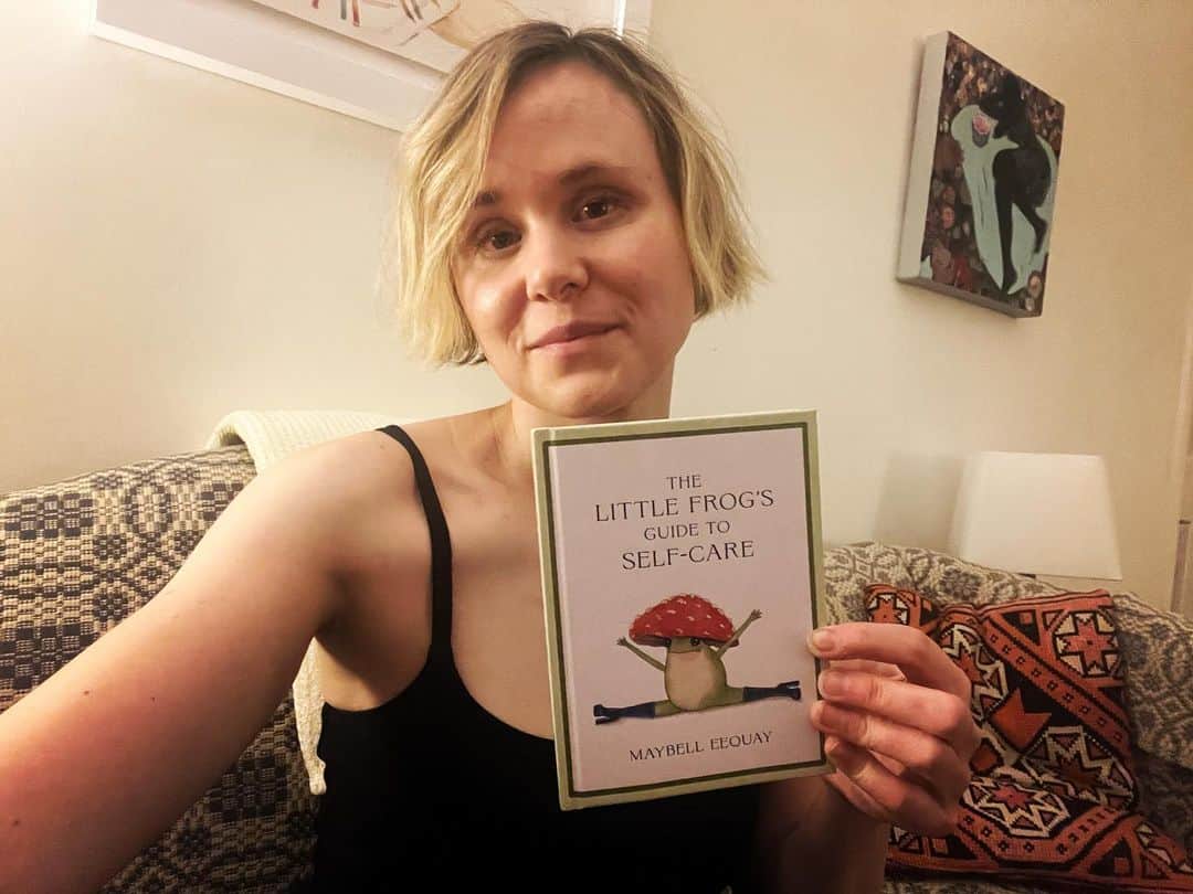 アリソン・ピルのインスタグラム：「Oh my goodness it’s here!  @maybell.eequay has written the most wonderful book starring Little Frog!  It’s full of wisdom, and feels like a soul hug.  If you or someone you know is just having a tiiiime, this book will pick you up. (Image description: a blonde white woman in black tank on blue and white sofa holds a book called The Little Frog’s Guide to Self-Care)」