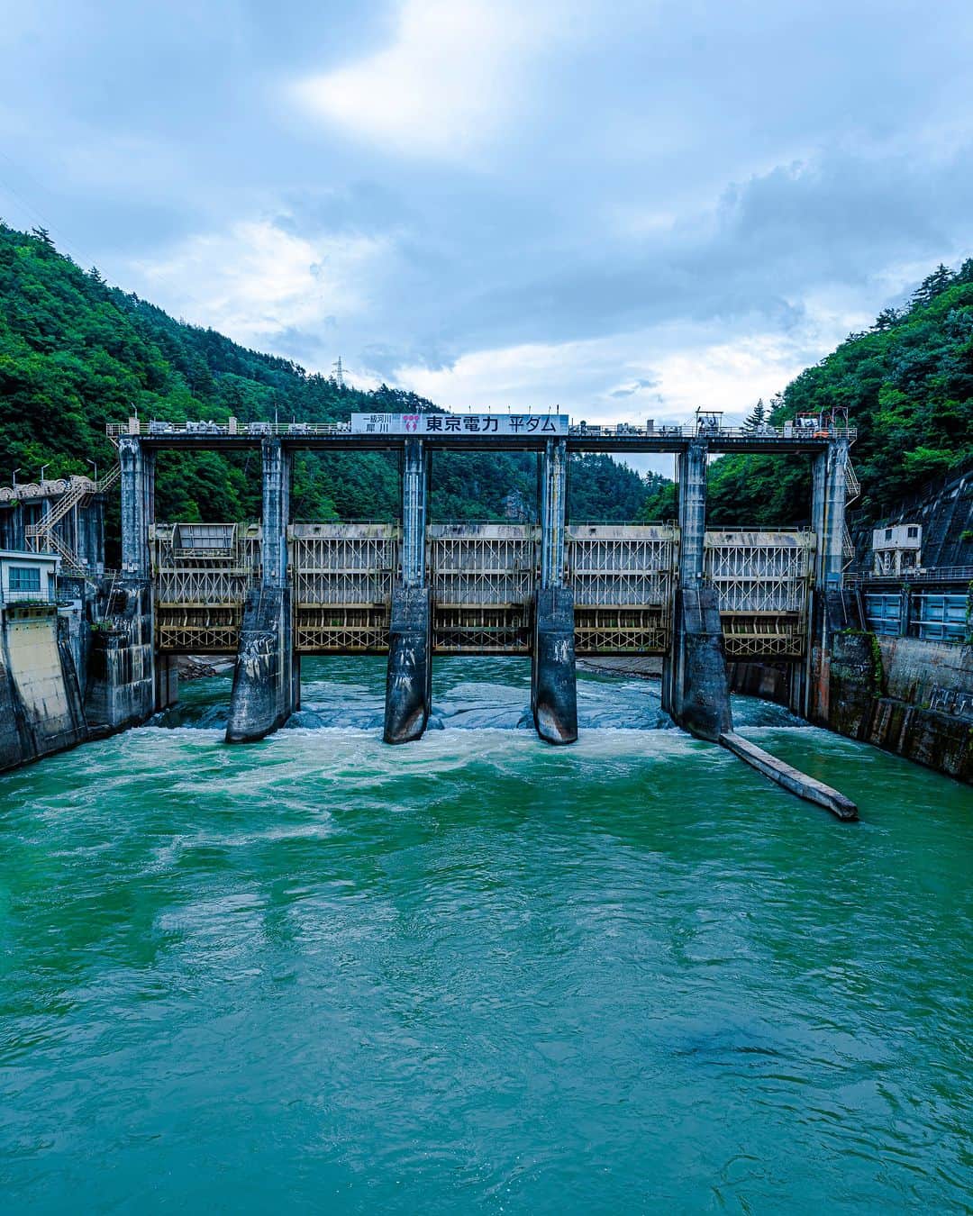 東京電力ホールディングス株式会社のインスタグラム：「平ダム Taira Dam  平ダムは、長野県長野市にある重力式コンクリートダムです。 洪水吐ゲートにはローラーゲートを採用しています。通常はゲートを閉めた状態で、河川から発電用に取水しています。貯水容量を超えないよう、降雨等による河川増水時は取水を停止して、全てのゲートを開き、ダムに貯まっている水を放流し自然河川の状態にします。 ダム上流は山清路（さんせいじ）と呼ばれる名勝で、穏やかな川の流れに険しい山々が映えます。  #東京電力 #tepco #長野県 #ダム #ダム巡り #ダム好き #damphotography #damphoto #水力発電 #再生可能エネルギー #renewableenergy #renewable #renewablepower #夏 #風景写真 #景色写真」