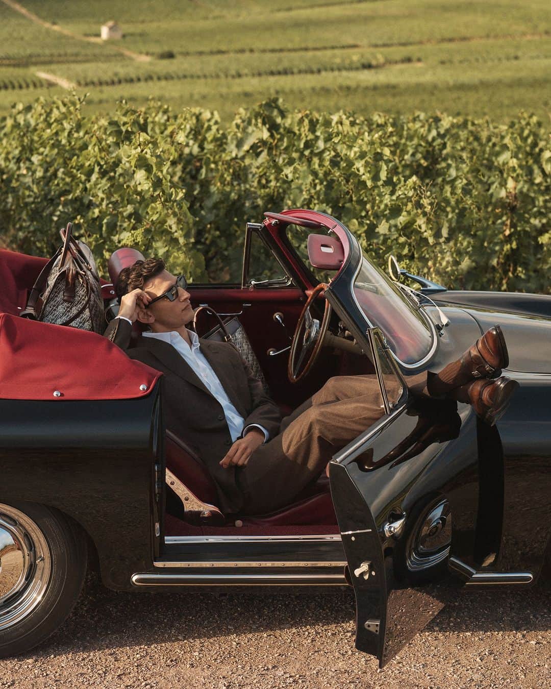 ベルルッティのインスタグラム：「- TRAVEL ICONIC: TOILE MARBEUF BY #BERLUTI -   An effortlessly chic suit. A vintage car with an eccentric twist. A dramatic luggage set: the Berluti man hits the road away from the city skyline and towards the glowing vineyards.   It’s all about the journey as you take a pit stop to smell the fragrant vineyard air, lazily basking in the sunlight in a nonchalant wool suit and Galet shoes, savoring every moment. The destination is almost an afterthought when you travel the Berluti way.   Now available online and in-store.  For the occasion, Berluti unveils an original Porsche 356 Cabriolet, customised with Saint-Émilion patinated Venezia leather seats, a baggage rack for unique road trips.  This story takes place at the Château de Saran, at the heart of the @moetchandon estate.   Creative direction by @trendsandculture for Berluti​ Styling by @benmartinengo  Photo by @danielrieraofficial  Model: @thibaudcharon ​  #BERLUTITOILEMARBEUF  The technical aspects of the car include the Flat 4-cylinder petrol engine, 1,582 cm3 displacement, 60 horse power, and twin carburetors.」