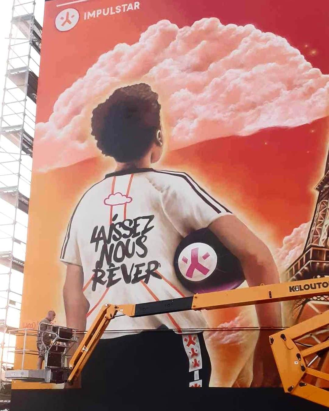 猫沢エミさんのインスタグラム写真 - (猫沢エミInstagram)「ではリクエストにお応えして、彼 @yannlazoo の最近の壁画作品もちょっとご紹介しますね。  こちら、フランスの若手スポーツ才能発掘・支援をしている大きな組織 @impulstar のPR壁画として依頼があった、大志を抱く少年とエッフェル塔🗼  あんまり壁がデカすぎると、逆に大きさがつかめなくなりますが、2枚目のクレーンと比べて、その大きさがわかるでしょうか。  A4サイズの元デッサンを、比率そのままにパースを狂わせず拡大させる技術は毎回圧巻（ヤンヤンの頭の中、どーなってんの？的な😵‍💫）。  しかも今回の依頼は、夕焼けのグラデーションがそのままアンプリュ・スターのシンボルカラーとリンクしているため、厳密な色の再現が求められた。  作画のための画材予算出しでも、一平方面積あたりのインクの消費量を細かく計算して、必要な数のスプレー缶量を出すなど、緻密な下準備が続いた。  この人は、右脳と左脳が両方バランスよく働いているタイプのアーティストだなと思う。  ここまで大きな壁画の場合、櫓を組んでも効率が悪いため、クレーン操縦士との二人三脚のオペレーションが必要になる。これだと、下まで降りずに、ぐーんと壁から離れてもらえれば全体を把握できるからいいんですって。  アーティスト友達にひとりアシスタントに入ってもらって、5日ほどで完成。  「俺もこんな体力勝負な現場は、あと何年できるかって感じだな〜」と言いながらも、彼の出発点であるストリートでのライブな仕事期間は、顔が少年に戻るよね👦  #猫パリ　#猫沢エミのインターナショナル五十路ライフシフト  #猫沢エミリーパリへ行く  #猫沢_art paris」9月8日 23時53分 - necozawaemi
