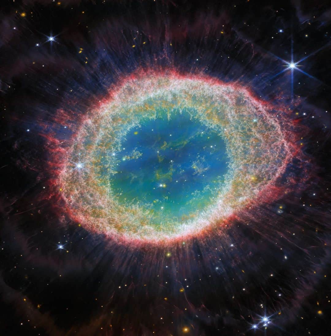 NASAのインスタグラム：「Name: Ring Nebula 🌟  Type: Planetary nebula, or an expanding shell of glowing gas expelled by a star late in its life.  Relationship Status: Possibly taken? @NASAWebb data may help us find out if the star at the center of this nebula may have a companion!  Learn more about this nebula at the link in our bio!  Image description: This image of the Ring Nebula, taken by Webb’s Near-Infrared Camera, appears as a distorted doughnut. The nebula’s inner cavity hosts shades of blue and green, while the detailed ring transitions through shades of orange in the inner regions and pink in the outer region. The ring’s inner region has distinct filament elements. Stars litter the scene, with a particularly prominent star with 8 long spikes in the top right corner.」