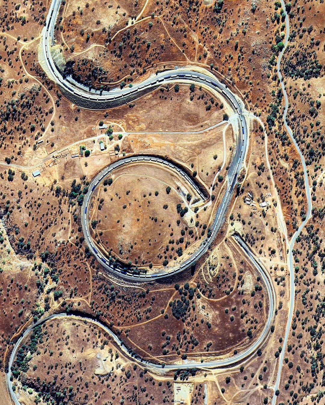 Daily Overviewさんのインスタグラム写真 - (Daily OverviewInstagram)「A train passes through the Tehachapi Loop — a 3,779-foot-long (1.15 km) spiral of railroad running through the Tehachapi Mountains in southern California. Constructed between 1874-76, the track rises at a steady two-percent grade through the Tehachapi Pass, gaining 77 feet (23 m) in elevation over a 1,210-foot-diameter (370 m) circle. Any train longer than about 56 cars, like the one seen here, passes over itself when going around the loop.  Created by @benjaminrgrant  Source imagery: @maxartechnologies」9月3日 1時11分 - dailyoverview