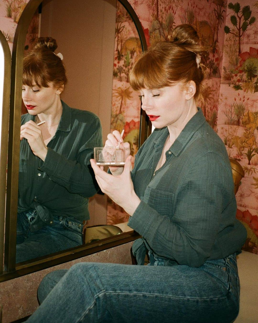 ブライス・ダラス・ハワードのインスタグラム：「💋💋💋⁣⁣⁣ ⁣ 📸: @andiejjane  ⁣⁣⁣ [ID: In blue jeans and a teal linen blouse, BDH sits on her vanity by the mirror. She wears her hair up in a clipped bun and applies Bakeup Beauty’s neutral palm eyeshadow palette and Yolaine’s red lip.]」