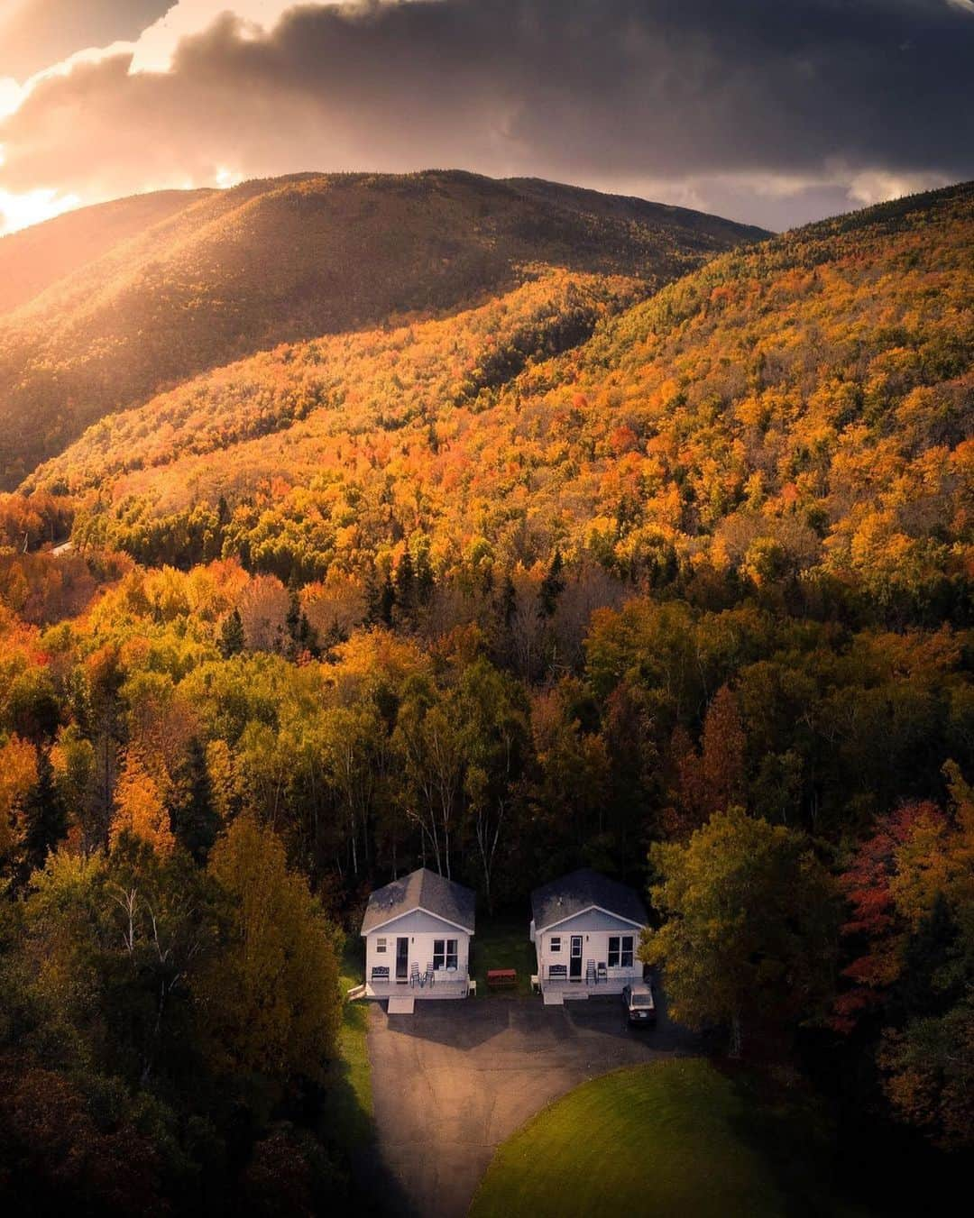 Explore Canadaのインスタグラム：「When you and your bestie get all cozied up for fall. 🍁     📷:@justsayantan 📍: @visitcapebretonisland, @visitnovascotia   #VisitCapeBreton #VisitNovaScotia #ExploreCanada   Image description: A photo of two white houses sitting side by side in front of a forested mountain whose leaves are a mix of yellow, orange, and green hues. A golden ray of sun lights the corner of the mountains.」