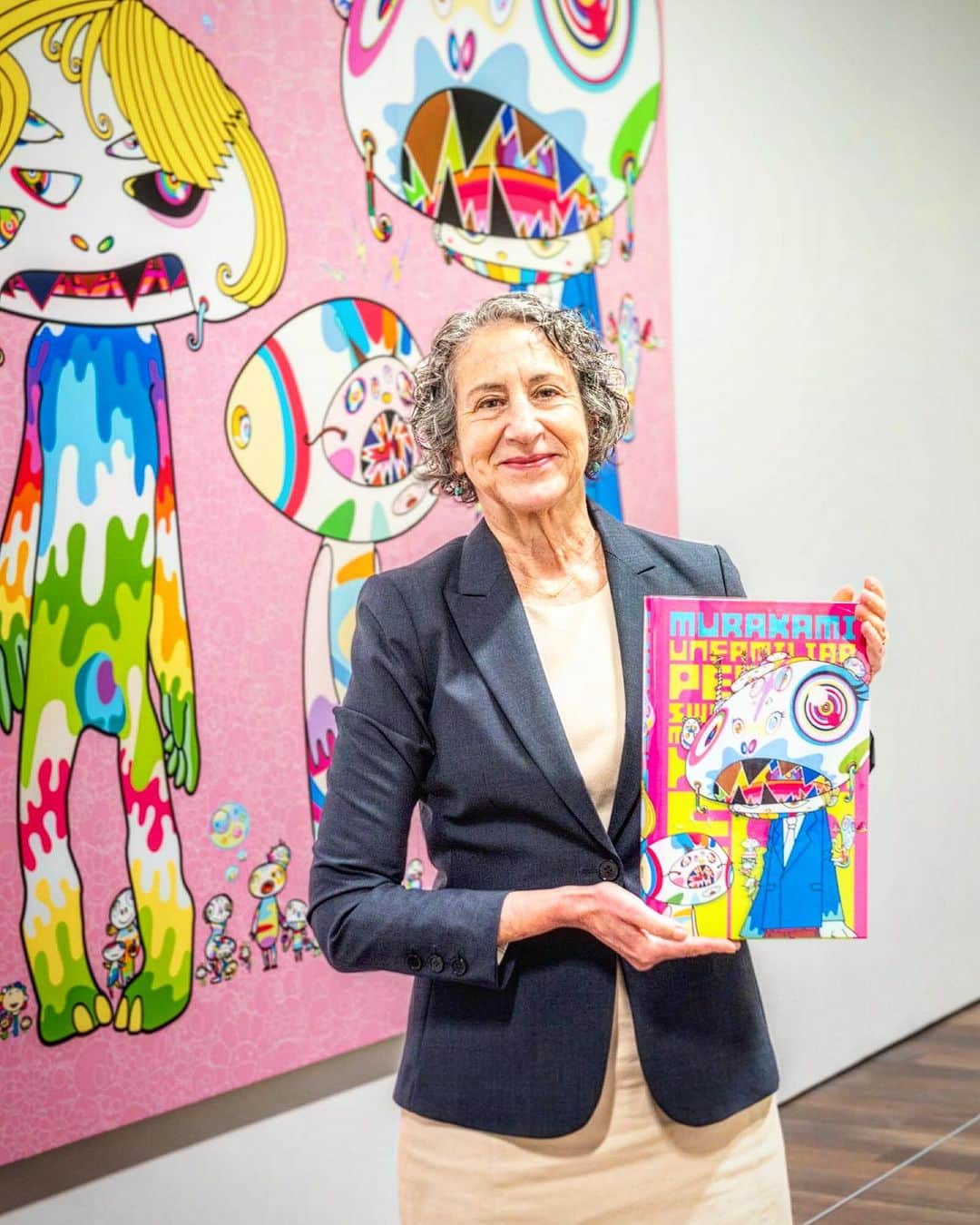 村上隆のインスタグラム：「My exhibition "Murakami: Monsterized" is now open at the Asian Art Museum in San Francisco @asianartmuseum . The photo is of the curator, Laura Allen, with the catalog for the exhibition, which she wrote and edited. The first print run was 10,000 copies, and two days before the exhibition opened, additional printing was already decided. The second print run will be 9,500 copies, I am told. There are several approaches to making an exhibition catalog. One way is to include the photographs of the actual installation on site and focus on documenting the exhibition. This time, the approach was a little different, and the catalogue is aimed at the visitors to communicate the concept of the exhibition. There is a painting series titled “Unfamiliar People" that I started to create during the pandemic, and the imagery of the first painting really seemed to resonate with Laura, so she selected it for the cover. In my own surrealistic way, I portrayed in this work the changing state of people who become skeptical and uncertain due to the pandemic. This is my first exhibition at a museum in San Francisco, and it seems that expectations were very high; I am told that the catalog is flying off the shelves. I hope that you will get ahold of the the catalog, whether in person at the museum or through online shops, and experience the realistic sense of how we create an exhibition.」