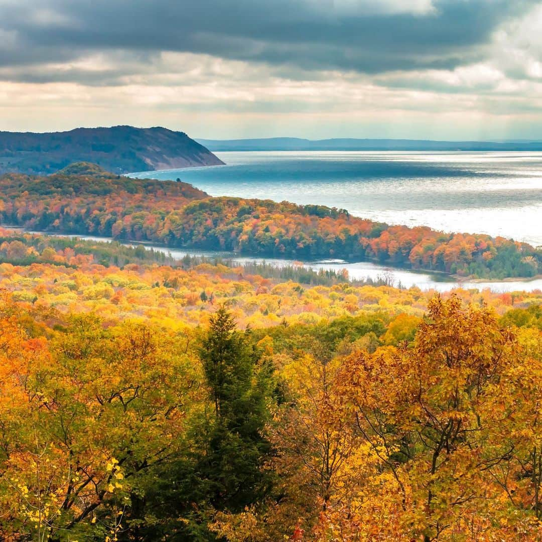 アメリカ内務省のインスタグラム：「We love the sweeping views of Lake Michigan and the beautiful fall colors that @SleepingBearNPS' sky-high dunes affords during this time of year. The park is diverse, embracing quiet, birch-lined streams, dense beech-maple forests and rugged bluffs towering above Lake Michigan.    So, come make lifelong memories, take a hike, have fun and develop a deep connection to this special place.    Photos by Nathaniel Gonzales    #sleepingbeardunes #michigan #publiclands #lakemichigan #fall   Alt Text:    Photo 1: A forest showing bright fall colors slopes down a hill to a blue lake during a partly cloudy day.    Photo 2: Mixed terrain of towering sand dunes and vibrant autumn foliage.」