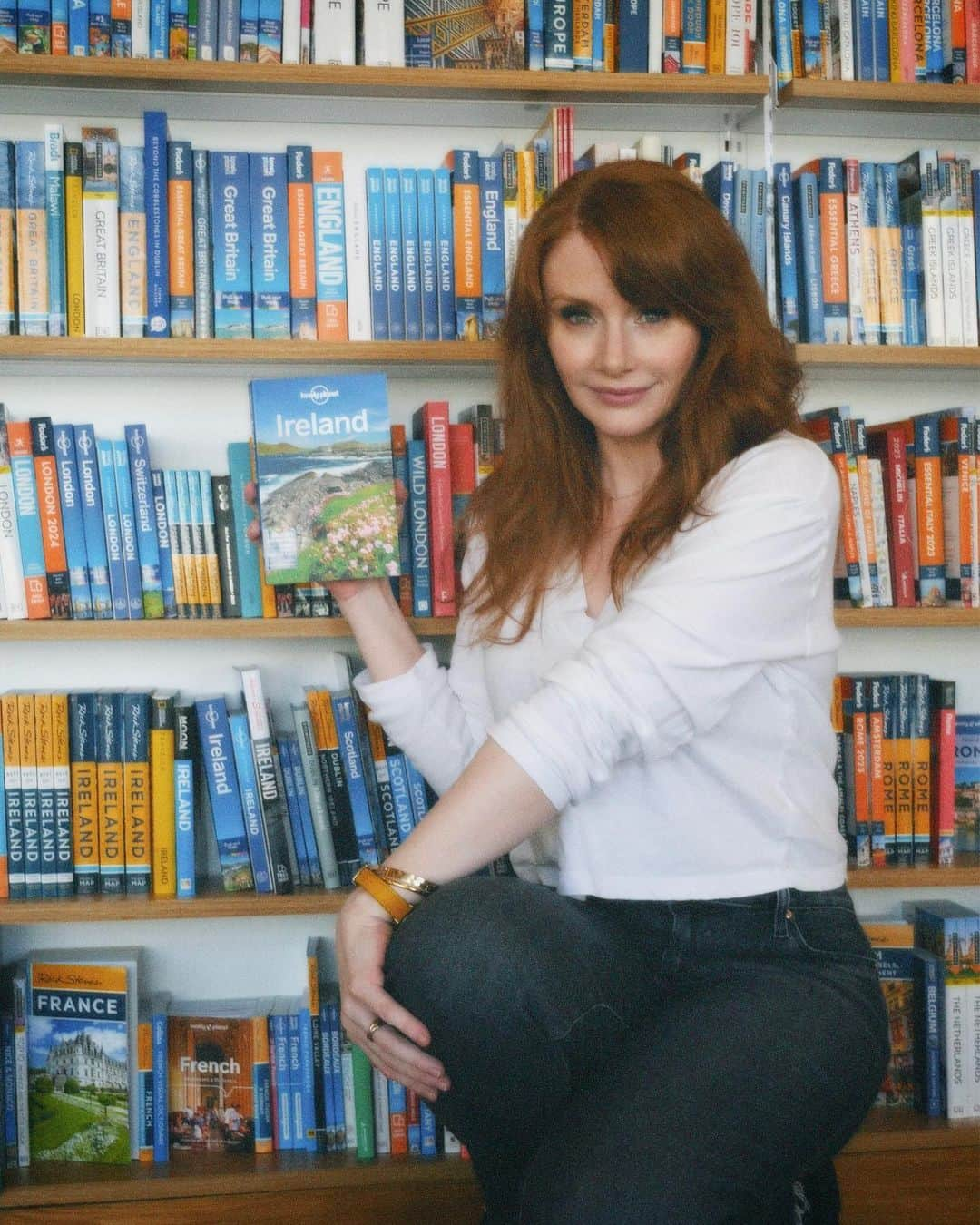 ブライス・ダラス・ハワードのインスタグラム：「Returning to the motherland this week #Ireland 🇮🇪⁣⁣ ⁣⁣ 📸: @andiejjane⁣⁣ ⁣⁣ [ID: Wearing a white blouse and blue jeans, BDH stands in front of a bookcase with shelves and shelves of miscellaneous travel guides. She holds the Lonely Planet guide for Ireland by her face, absolutely ecstatic about returning to Ireland after 30 years!!]」