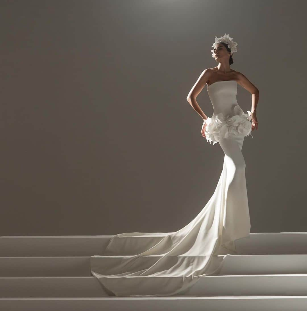 ステファンローランのインスタグラム：「RENDEZ VOUS Strapless mermaid-style long white crepe dress adorned with gardenias on the hips. White gardenia headpiece.  For this new Wedding collection, Stéphane Rolland continues to reinterpret the emblematic Haute Couture designs of the Parisian house.  Evolving two-piece dresses, asymmetrical tunics, draped ziberline bust sculptures, silk taffeta cascading skirts with trains, and crystal-embroidered cape dresses.  Silk-embroidered bouquets of gardenias adorn the headpieces and grace the hips and hemlines of dresses...   #StephaneRolland #Bridal #WeddingInspiration #NYBFW」