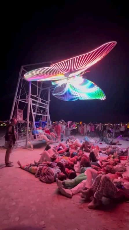 Padgramのインスタグラム：「Mariposa, a magnificent 26-foot LED butterfly sculpture created by the talented artist @Christopher.Schardt for Burning Man 2023, soars gracefully above the desert sands. Its wings possess a unique hinge mechanism that allows them to flutter, a whimsical dance orchestrated by adventurous visitors swinging on porch swings. Adorned with a mesmerizing array of over 39,000 LEDs, this radiant creation comes to life with the harmonious melodies of classical and classic pop music, accompanied by intricately choreographed visuals displayed on its luminescent canvas.  Crafted from sturdy aluminum tubing, Mariposa’s framework exudes elegance and resilience. LED-studded bars extend outward from its spine, illuminating the night with an enchanting glow. Each wing pivots gracefully around a front-to-back axis, adding a dynamic dimension to its majestic flight. Surrounding this enchanting butterfly, four speakers cleverly masquerade as colossal flower pots, their LED blossoms blooming in a symphony of light and sound.   Credits to @architectanddesign  • #pgdaily #pgstar#pgcounty #planetgo#planet #planetearth #amazing #awesome #art #design #burningman #burningman2023 #blackrockcity #mariposa #sculpture #butterfly」