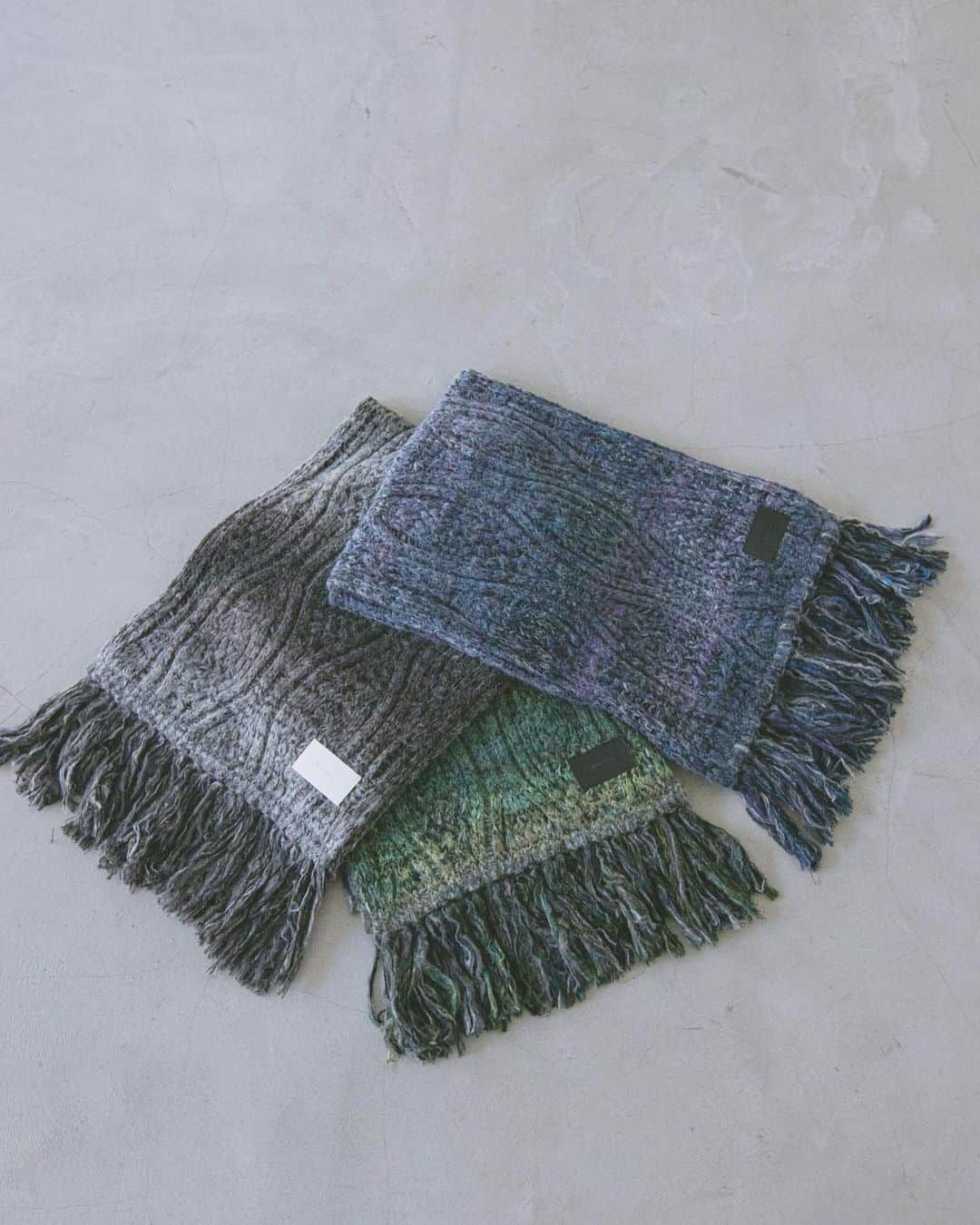 のインスタグラム：「. 2023 Fall / Winter Collection " FRAGILE "  Hazy knit Scarf  風に舞う粉雪。 霞んで見えたその光景の儚さと強さを絣糸を用いて表現。 配色の異なる2種類のかすり糸を用いる事で柄の表情に奥行きをもたせている。 1本1本丁寧に手作業でつけられたフリンジは靡く度にスタイリングにアクセントを生み出します。  ブランドネームが刻印されたレザーパッチとのコントラスト ざっくりと編まれたローゲージの質感 スタイリングのアクセントにも最適な逸品」