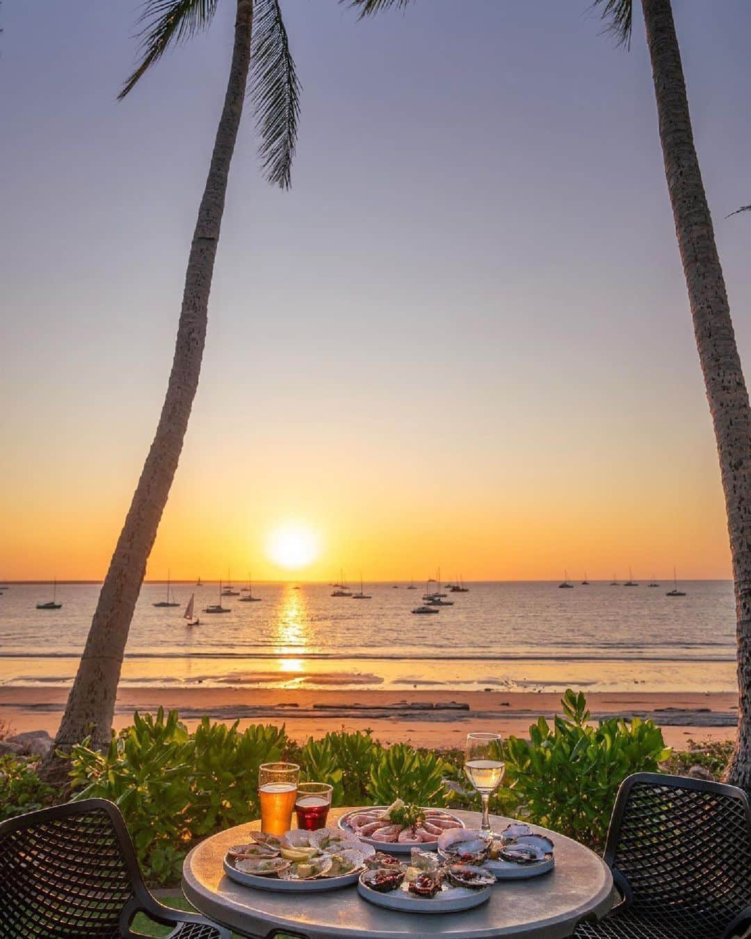 Australiaのインスタグラム：「Pull up a deckchair; we've saved you the best seat in the house! 🌞🍹 If balmy nights, colourful characters and outdoor adventures are on your #BucketList, you'll want to visit Gulumerrdgen (#Darwin) in @tourismtopend just like @_markfitz. After exploring the breathtaking national parks nearby (we're talking @litchfieldnationalpark, @seekakadu and #NitmilukNationalPark), sit back and relax at @thedarwinsailingclub for a drool-worthy seafood dinner while you watch the sun dip below the horizon.   #SeeAustralia #ComeAndSayGday #NTAustralia #TourismTopEnd  ID: a seafood spread and a few beverages are arranged on a small table set for two. Two palm trees frame the view from the table - a sandy beach at sunset. Sail boats are anchored just off the shore.」