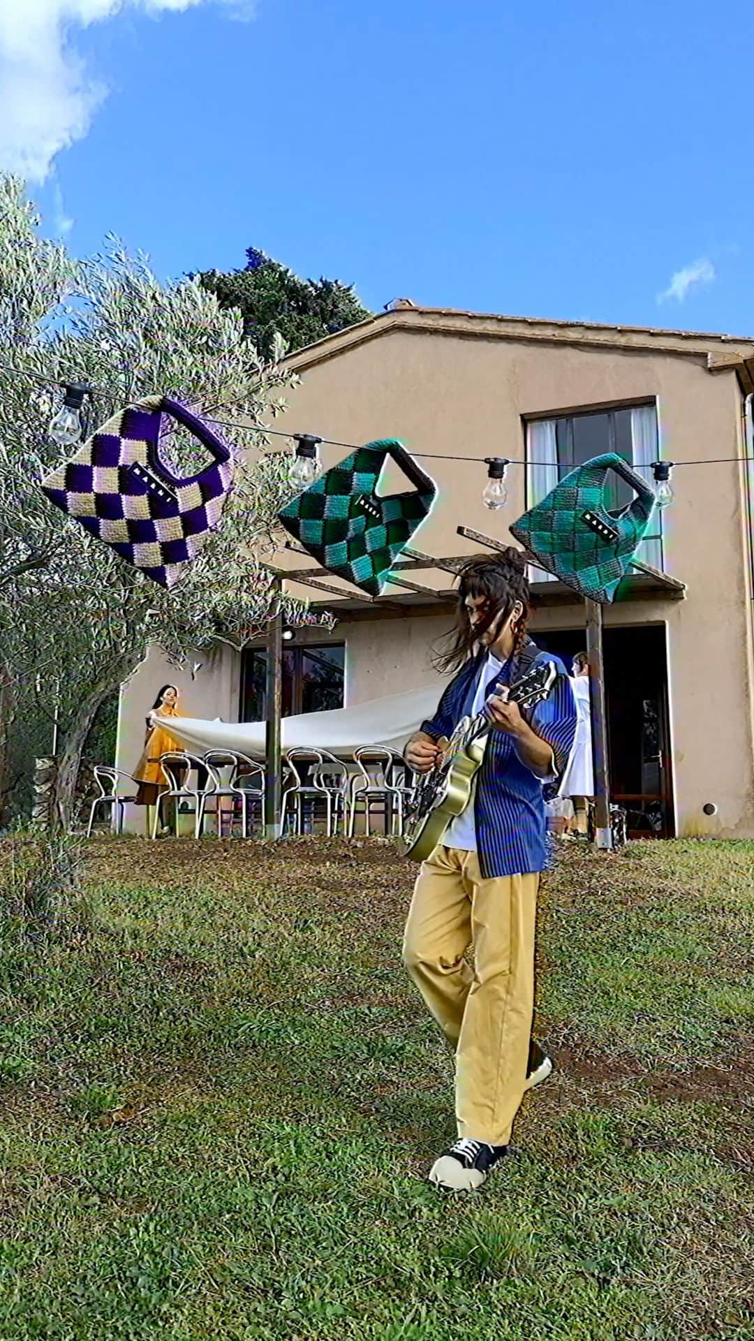 マルニのインスタグラム：「Danielle waits for friends for lunch and tunes his guitar to the wind. Above, there's laundry hanging, made of Crochet Chessboard Bags or woven Tech Wool Diamond Bags. Green, purple, blue. The color spectrum of Marni Market. Diamonds, checks, handcrafted patterns. With care.  The new #MarniMarket collection is now available online and in stores.  Video: @marcoproserpio_ Model: @danielle_musica  #marni」