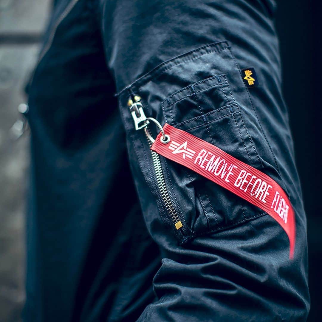 ALPHA INDUSTRIES JAPANさんのインスタグラム写真 - (ALPHA INDUSTRIES JAPANInstagram)「𝐑𝐄𝐃 𝐑𝐈𝐁𝐁𝐎𝐍  『REMOVE BEFORE FLIGHT※飛行前に取り外せ！』と記されたレッドリボン。MA-1などのシガレットポケットに配置されたこのスモールピースは2000年当初に採用されて以来、アルファ インダストリーズのシンボルとも称されている。そもそもこのレッドリボンは航空機が地上にいる際に、損傷してはいけない機構や装備などを固定し、誤作動を防止するための『グランドセフティーピン』の付属品がルーツ。整備員の抜き忘れ防止、更には引き抜いた際に操縦席へ向かって掲げることで、パイロットとピンが抜かれたことを共有しやすいように視認性を重視したデザインが特徴となっている。  𝐍𝐄𝐖 𝐒𝐄𝐍𝐒𝐄 𝐎𝐅 𝐖𝐀𝐋𝐔𝐄𝐒  冷戦中にコントラクターからミリタリーアパレルメーカーに転身を遂げたアルファ社だが、2000年以前はあくまでアメリカンカジュアルを好む男性向けのファッションブランドという側面が強かった。しかしながらその印象は、21世紀初頭に大きく覆される。各種大手メゾンが挙って軍衣料由来のコレクションを展開し、ミリタリーファッションが世界的に脚光を浴びる中で、リアルコントラクターとしてのルーツを持つアルファ社に注目が集まったのである。アルファ社はそのニーズにこたえるべく様々なミリタリーウェアを発売。その結果、レッドリボンがストリートに溢れることになった。 これこそが『武骨なミリタリージャケット』がオーセンティックな価値観を備えた『日常のアウター』へと変化を遂げた瞬間であった。   #alpha_industries_japan  #alpha_industries  #ALPHAINDUSTRIES #ALPHA #ALPHASHOP #MA1 #the_alpha_story #removebeforeflight #redribbon」11月22日 20時16分 - alpha_industries_japan