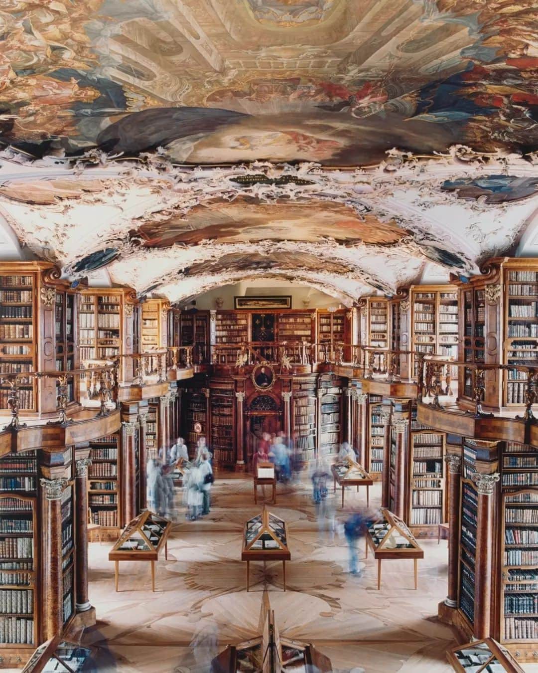 テート・ギャラリーのインスタグラム：「This magnificent photograph by German photographer Candida Höfer shows the interior of the Abbey Library of St. Gallen, the oldest monastery library in Switzerland. ⛪📚  Höfer is known for her large-scale colour photographs of impressive architectural spaces. Her spectacular images draw attention to symmetry, sense of scale, dramatic proportions and unlikely details. Her interiors are almost always empty of human presence. They place emphasis on silence and offer an opportunity for objects to project their voices. Unusually, this photograph includes images of people, but the long exposure time has rendered them blurry. Visitors to the library appear like ghostly apparitions, suggesting the transience of life and the passage of time.  You can find Höfer’s photograph in all of its splendour in our Tate Modern exhibition #CapturingTheMoment. Members go free. 🎟️  📷 Candida Höfer, Stiftsbibliothek St. Gallen I, 2001 © Candida Höfer」